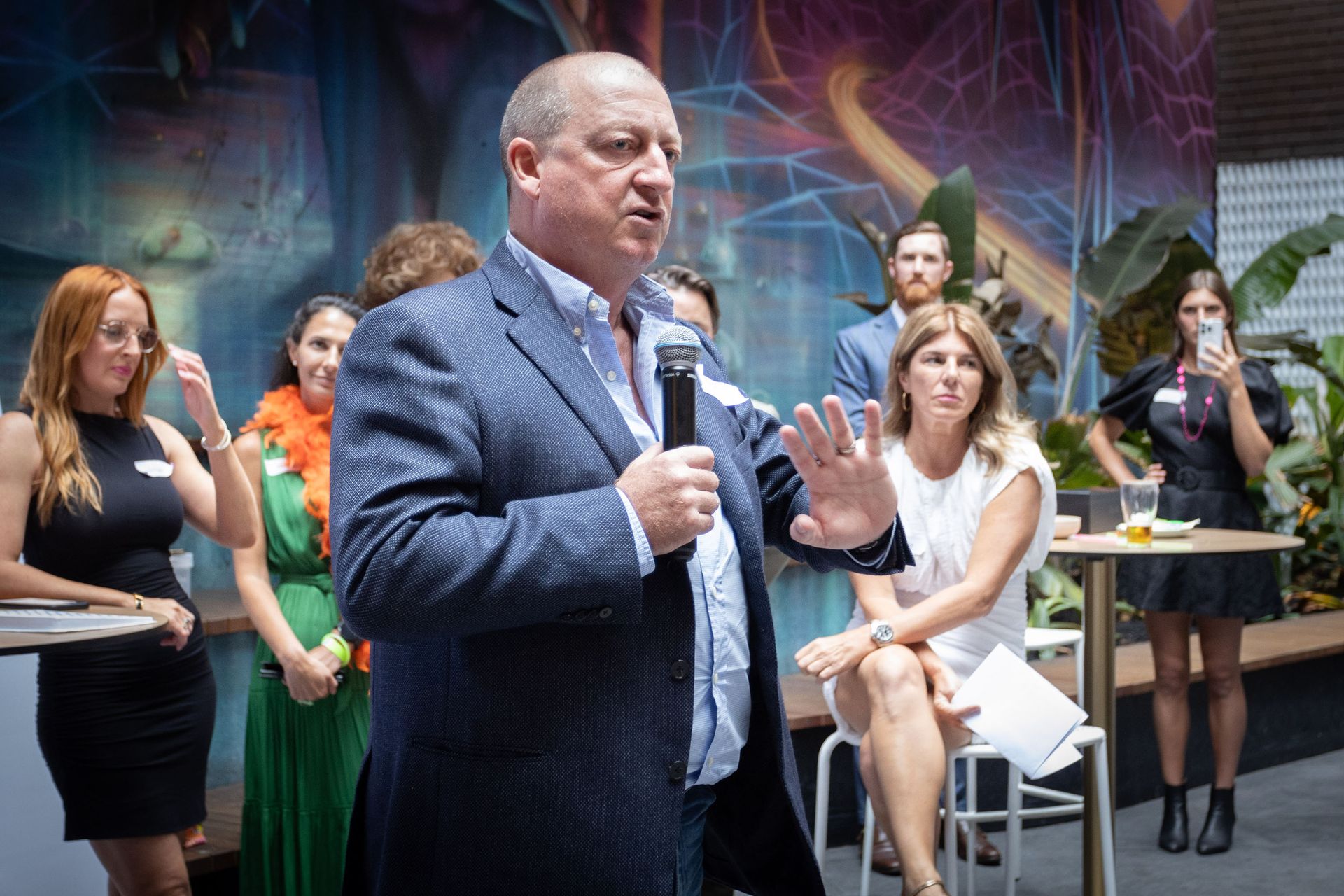 A person speaks into a microphone before a group of attendees in a bright, modern room with wall art and plants.