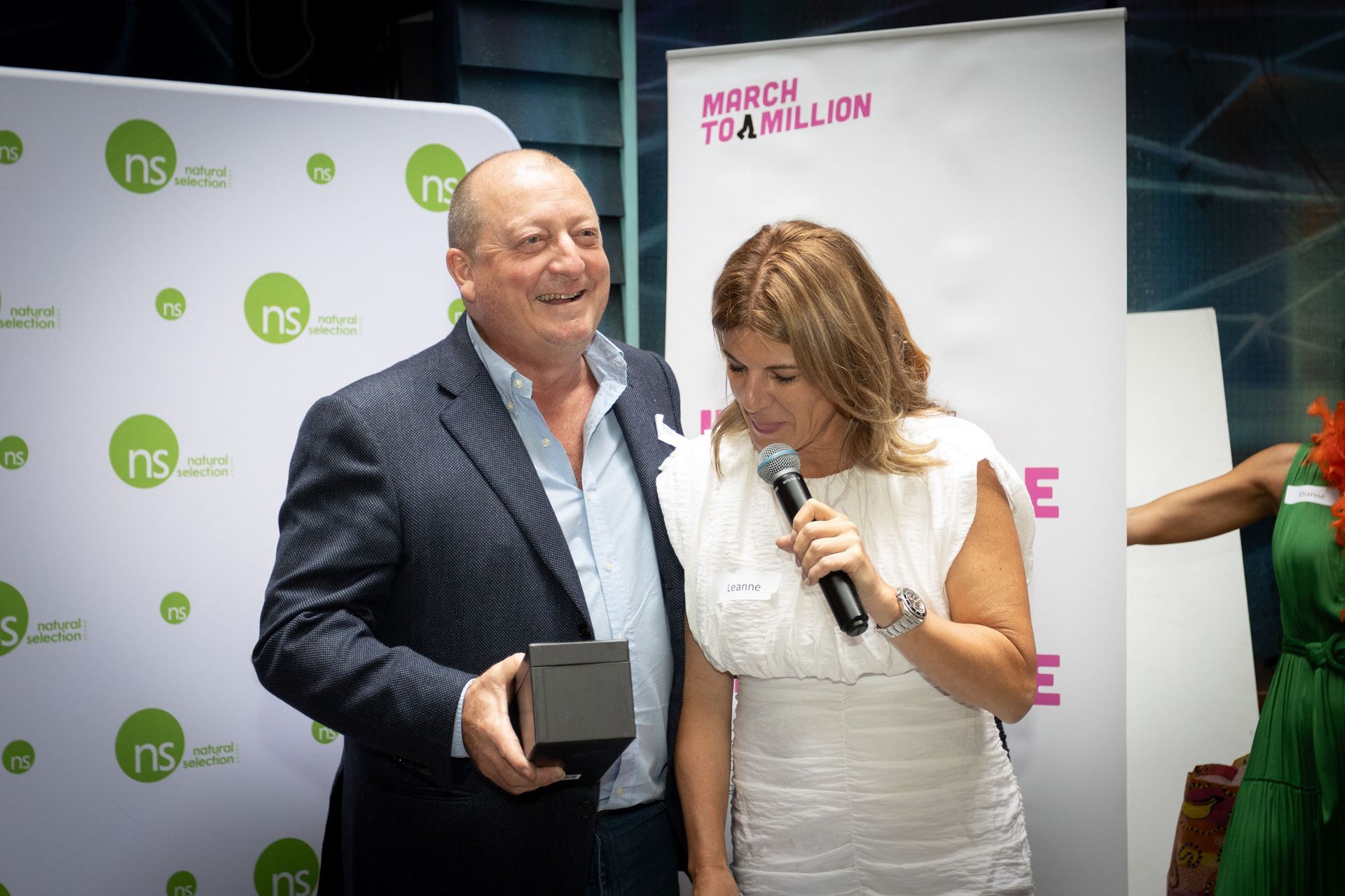 A man in a blazer holds an award while a woman speaks into a microphone at an event with 