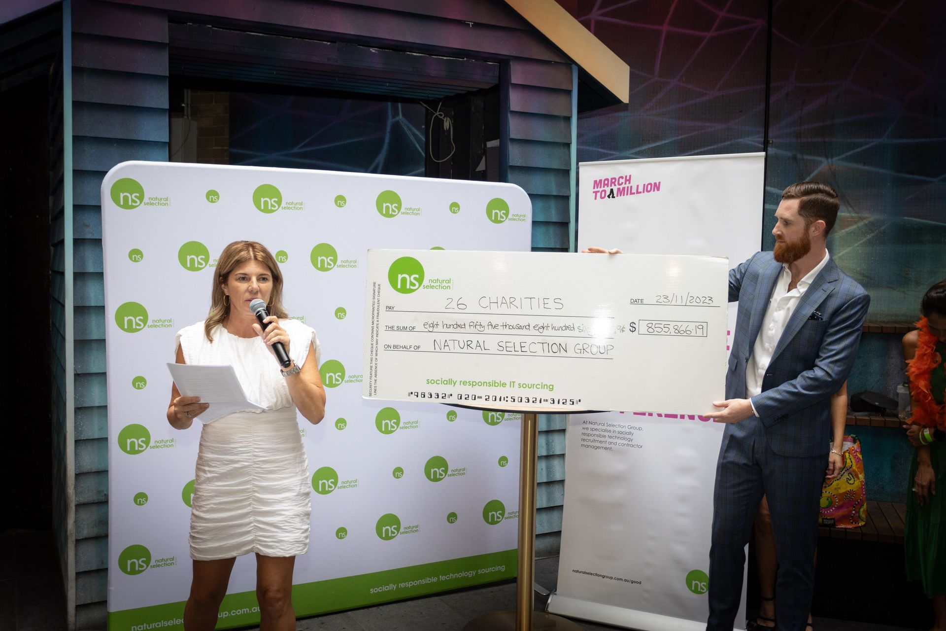 A woman with a microphone and a man in a suit stand by a large presentation check at an event with branded backdrops.