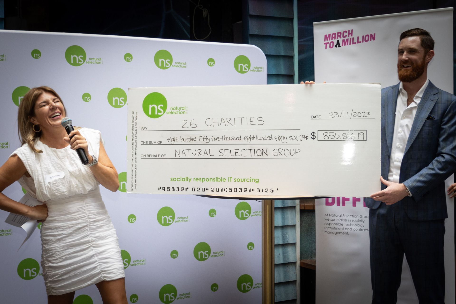 A woman and a man hold a large check representing a $333,333 donation from Natural Selection Group at a formal event.