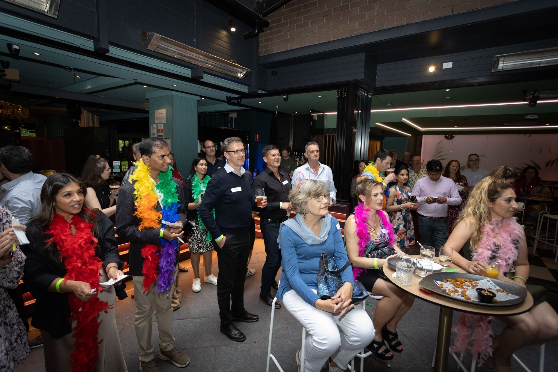 People in festive attire with colorful feather boas gather at a social event in a dimly lit, modern lounge.