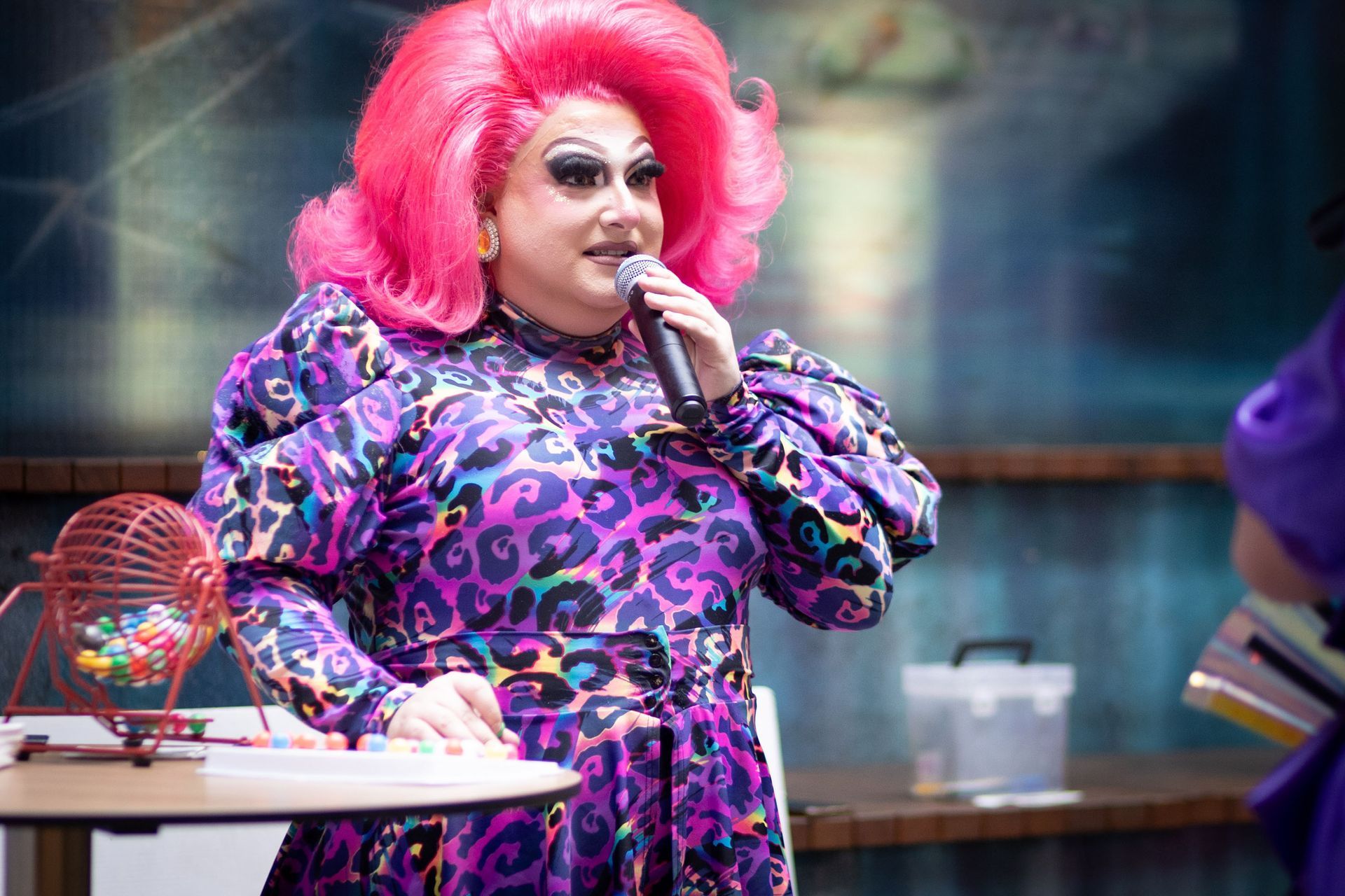 A performer with bright pink hair and colorful patterned clothing holds a microphone, standing beside a bingo cage.
