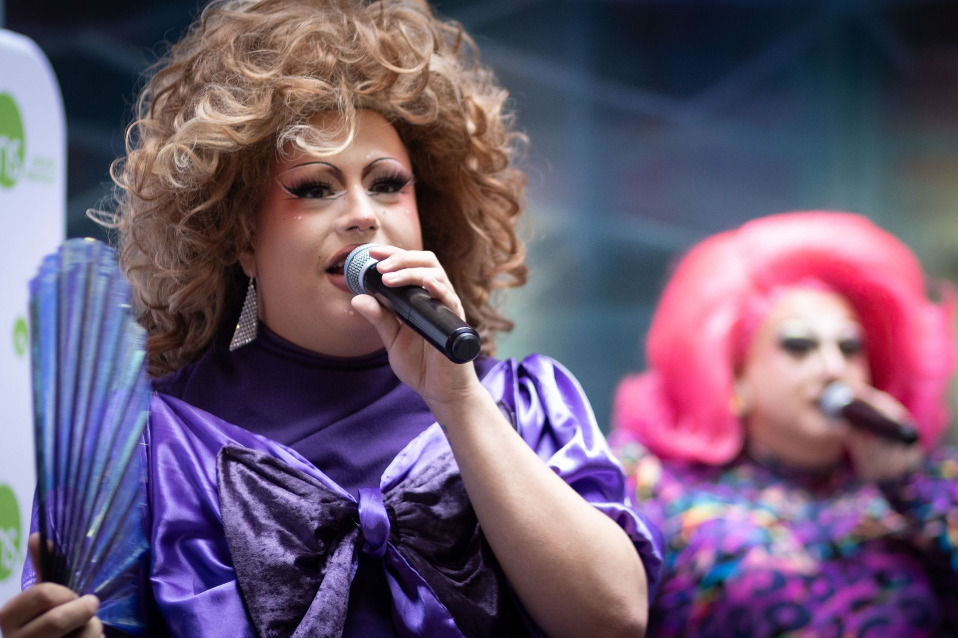 Two performers in drag, one with large brown hair and a purple outfit, hold microphones and address a crowd.