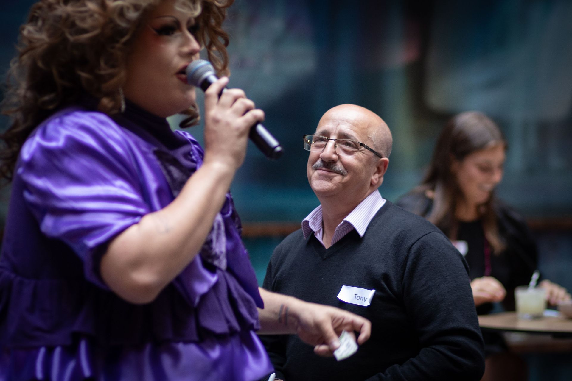 A person in a purple dress speaks into a microphone while a smiling person in a black sweater watches them.