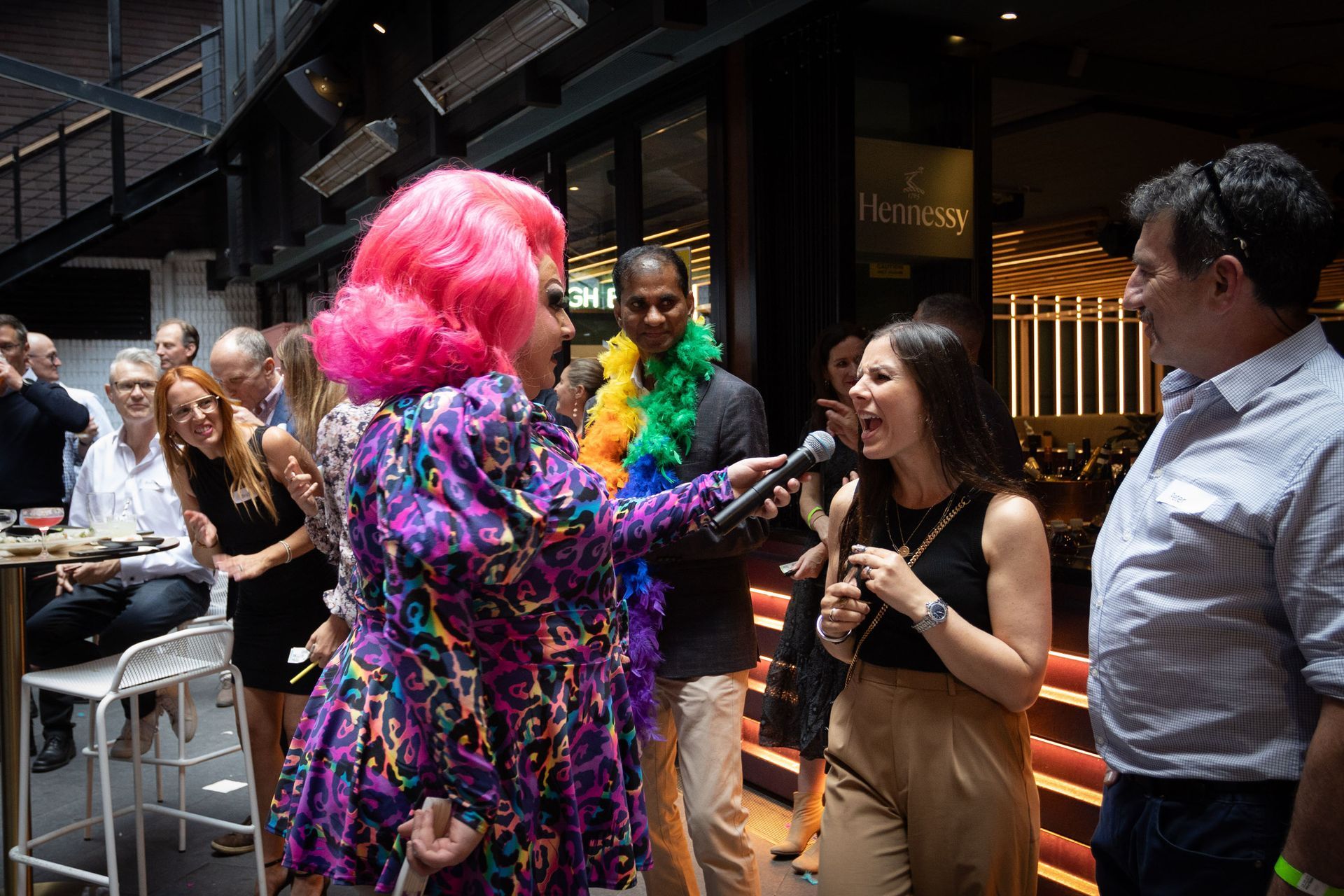 A person in a vibrant pink wig and patterned outfit holds a microphone for a woman, with others observing in a venue.