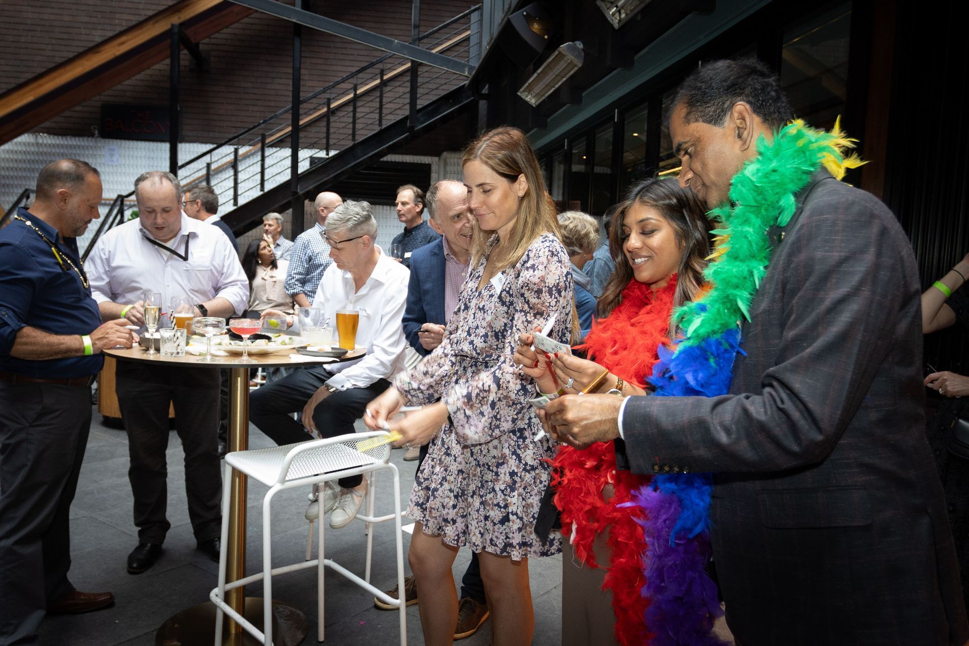 A group of people gathers at a high-top table in a modern venue, with some wearing colorful feather boas.