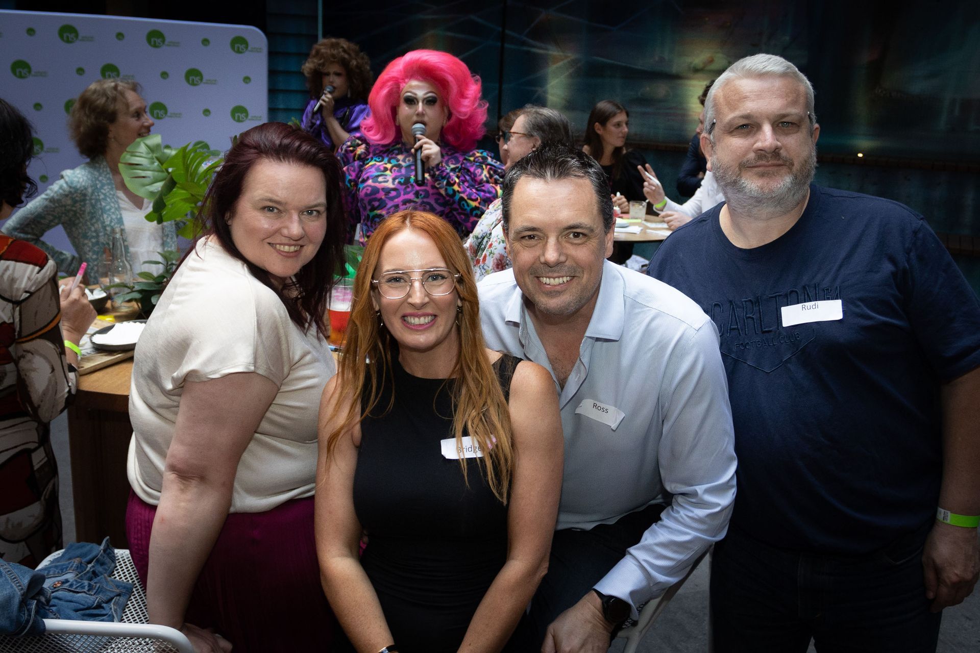 Four people smile for a group photo at an indoor event, with a performer in a bright pink wig visible in the background.