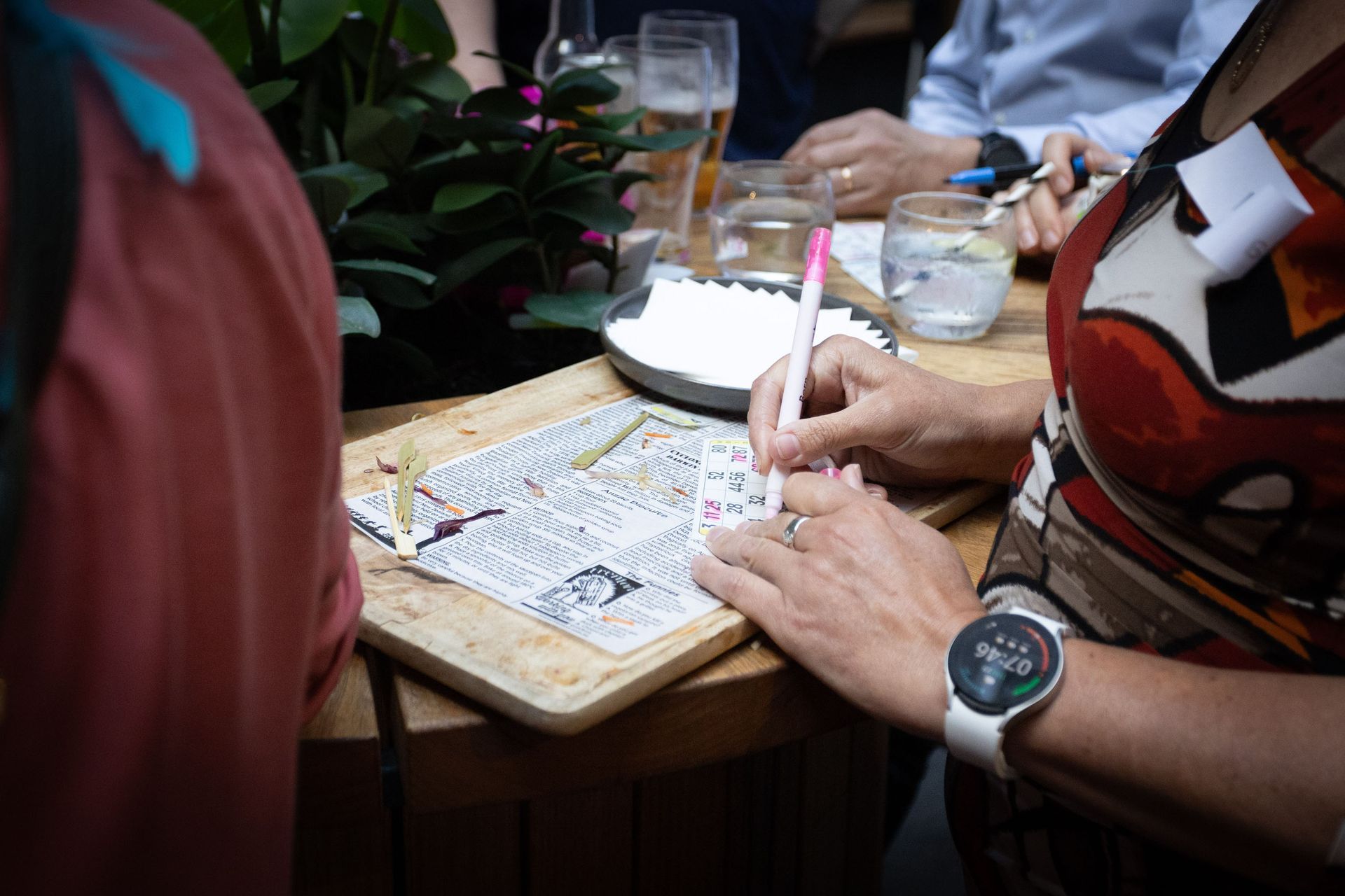 A person writes on a small paper at a wooden table in a social setting, with drinks and a plant in the background.