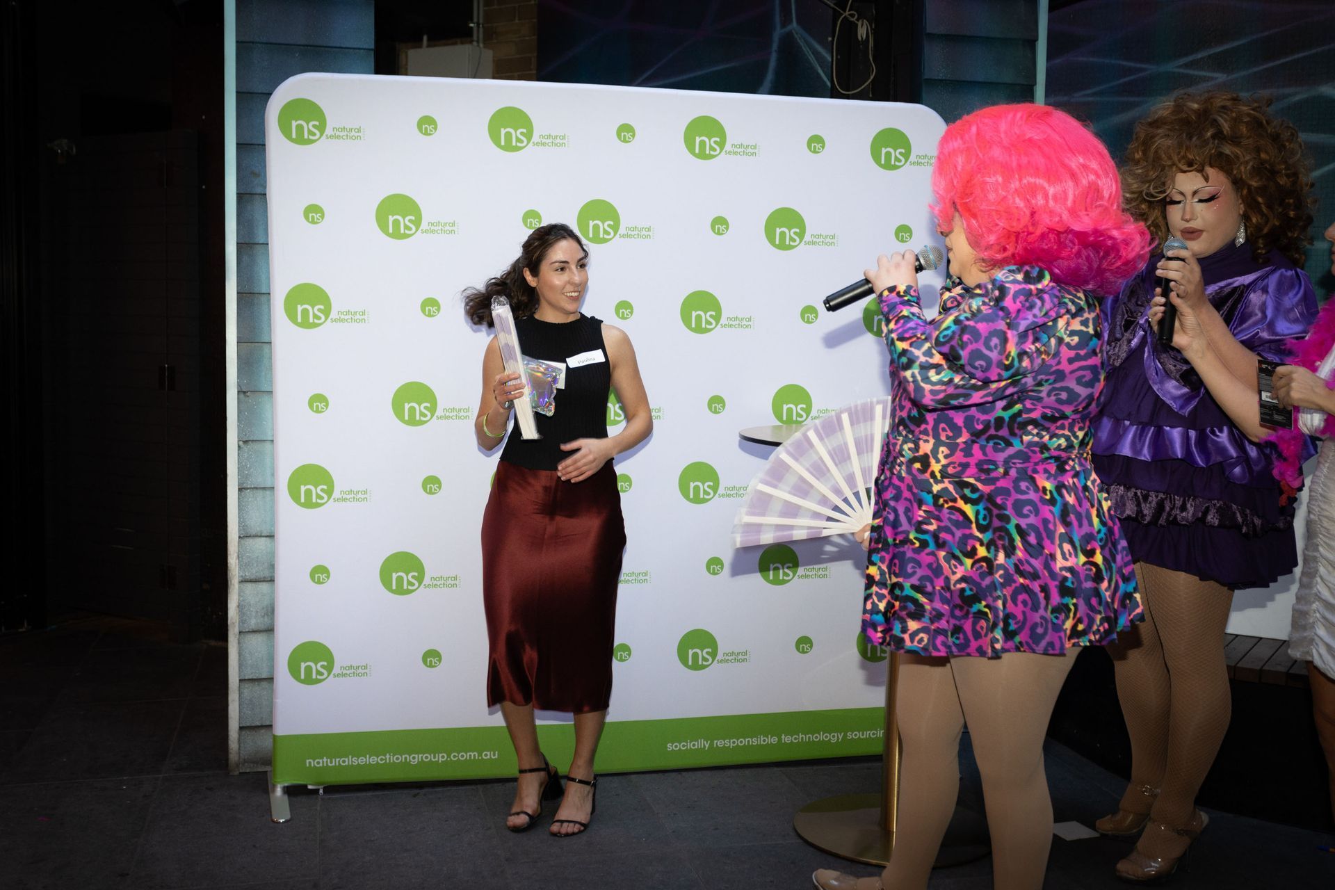 A woman holding an award stands on stage next to two performers with microphones in front of a branded backdrop.