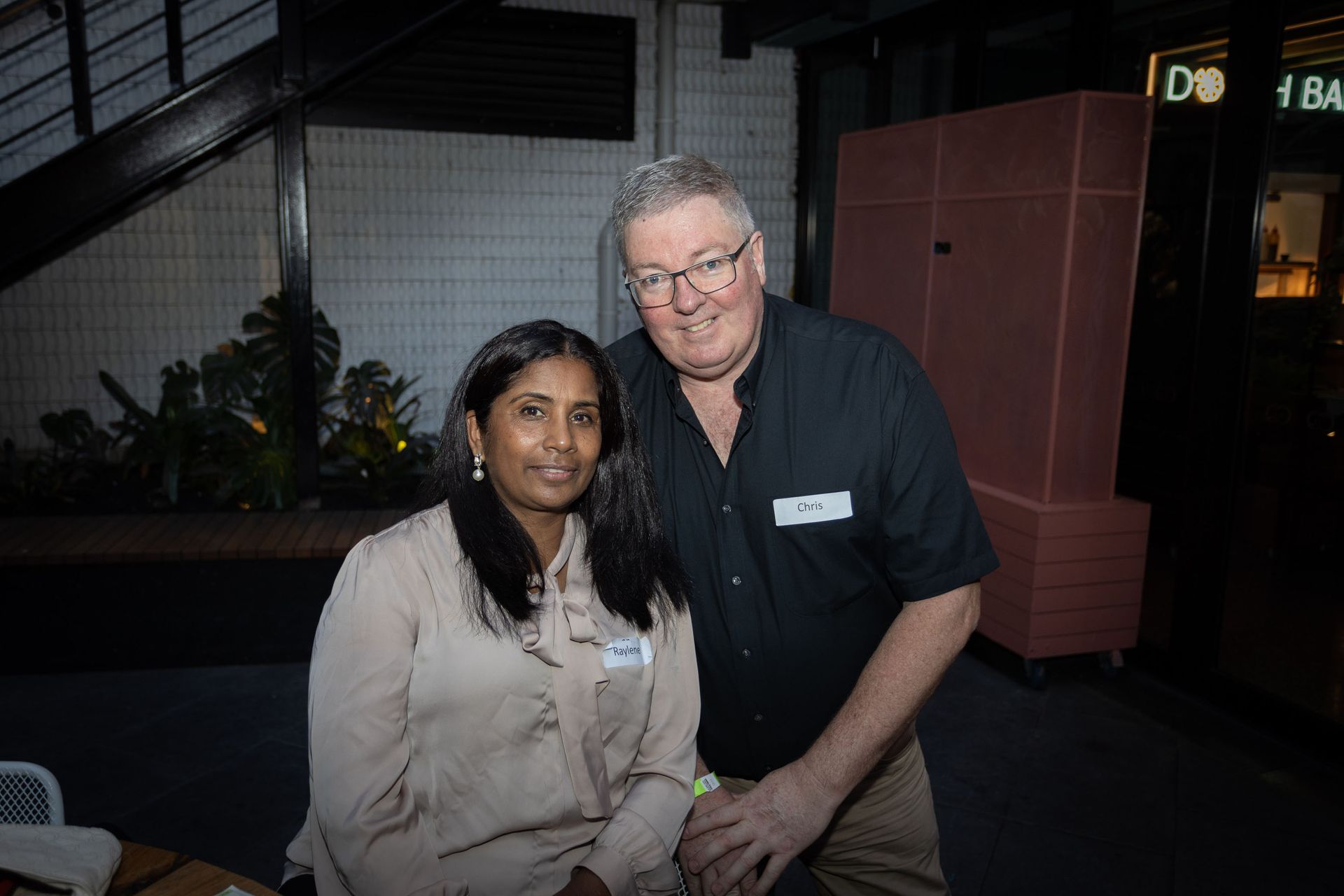 Two people posing for a photo indoors, both wearing name tags, with a dark background and an illuminated sign.