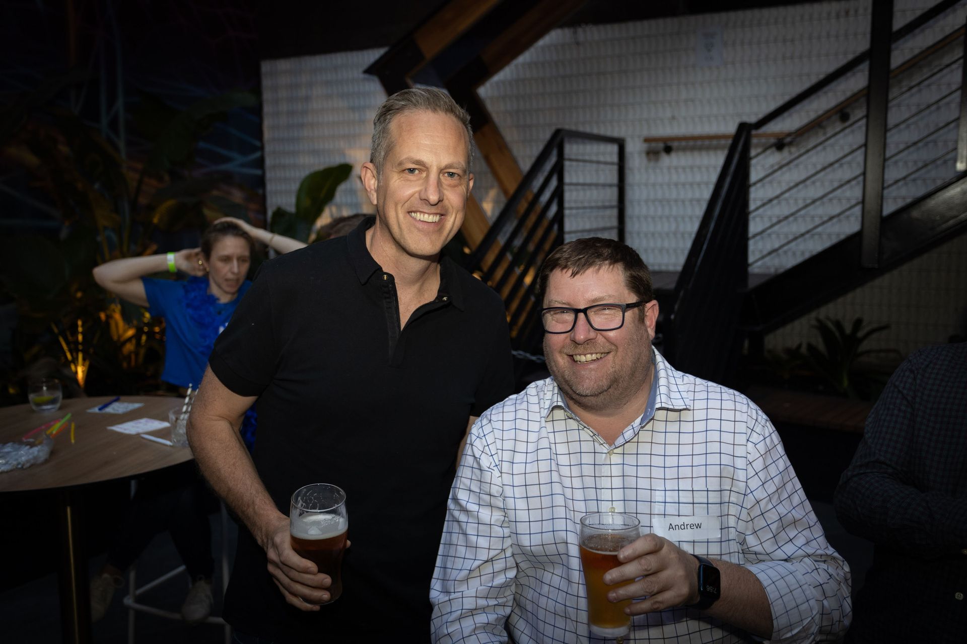 Two men stand smiling and holding drinks at an indoor event, with a woman in the background near a table.