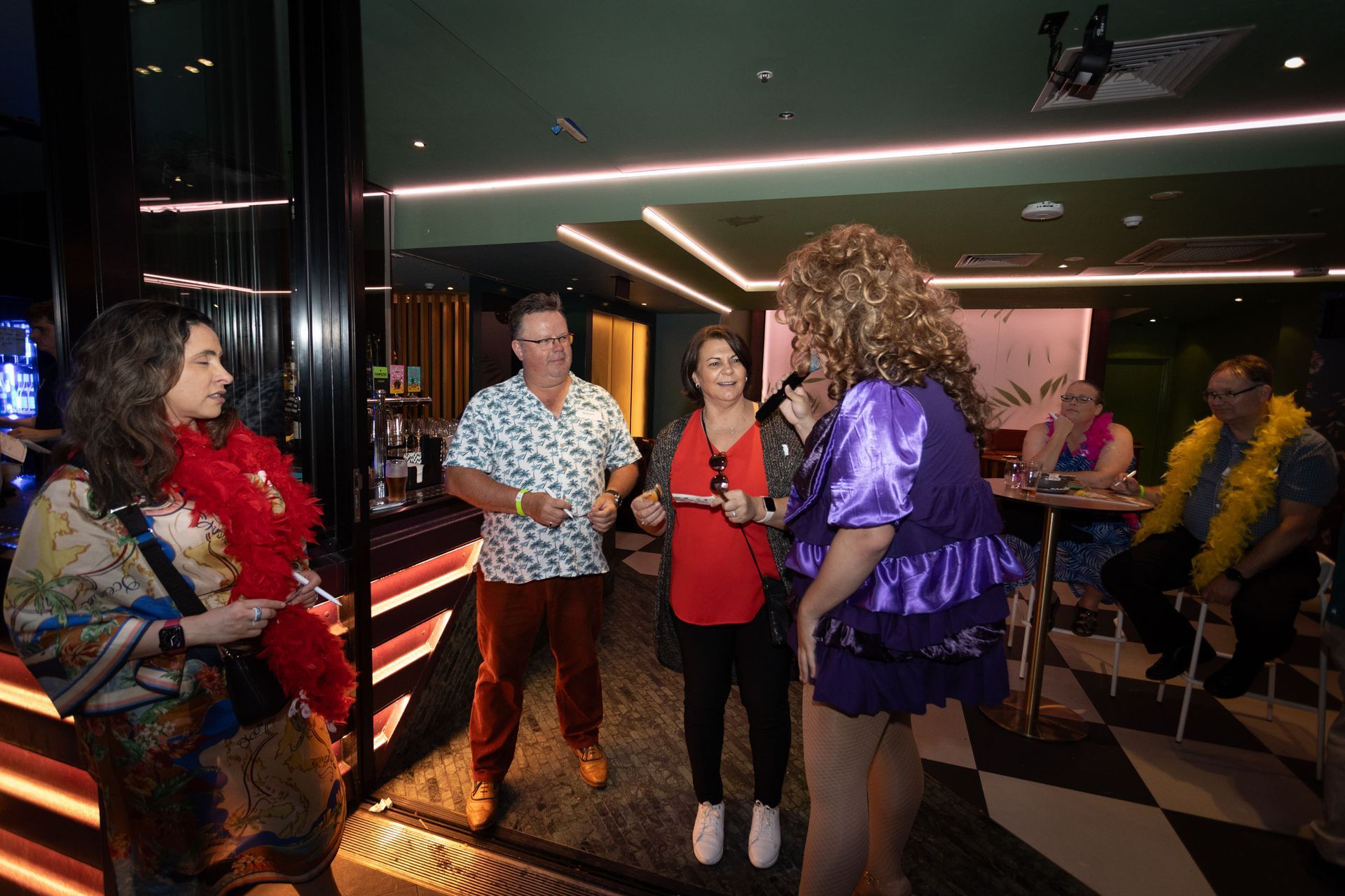 People socialize in a brightly lit bar, with a group standing in the center talking while others sit in the background.