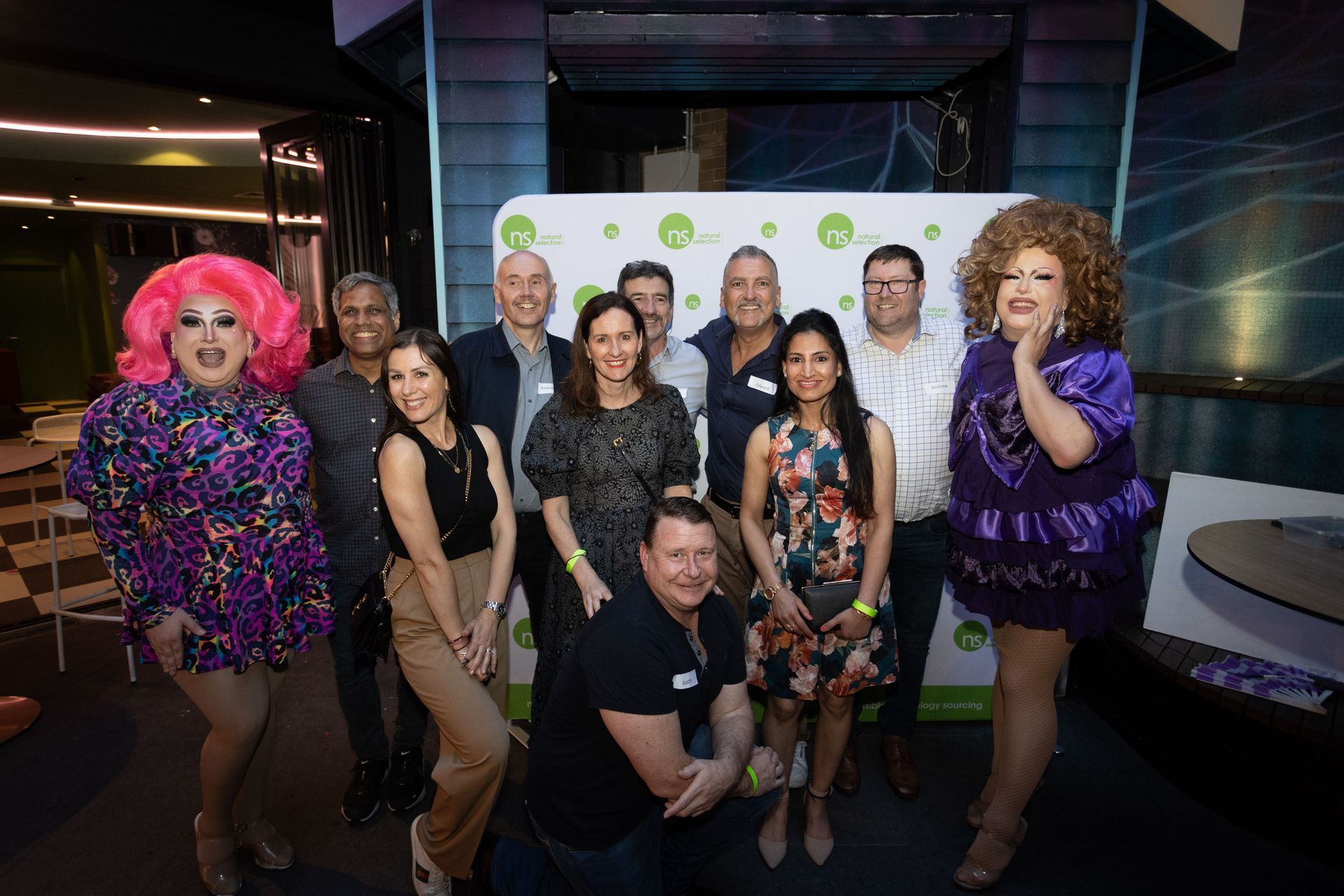 A diverse group poses for a photo in front of a white backdrop with circular logos at an indoor evening event.