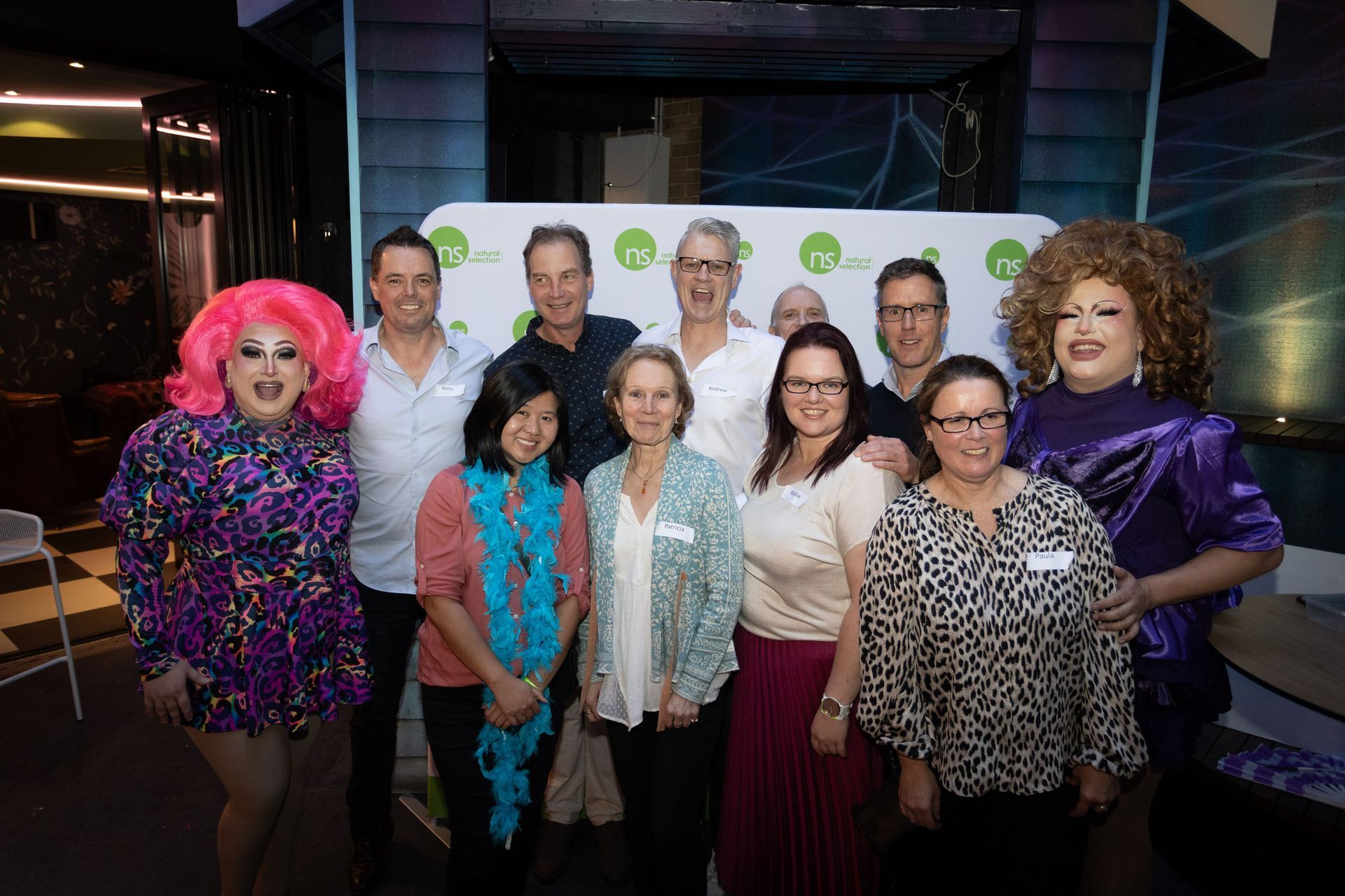 A diverse group of ten people stand in a line smiling for a photo in front of a white banner with green logos.