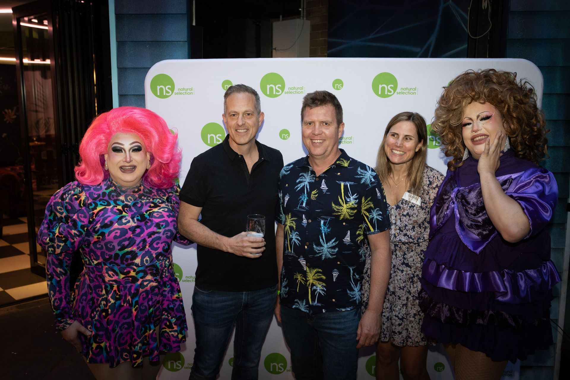 Two drag performers in vibrant outfits and two casually dressed individuals pose together in front of a branded backdrop.