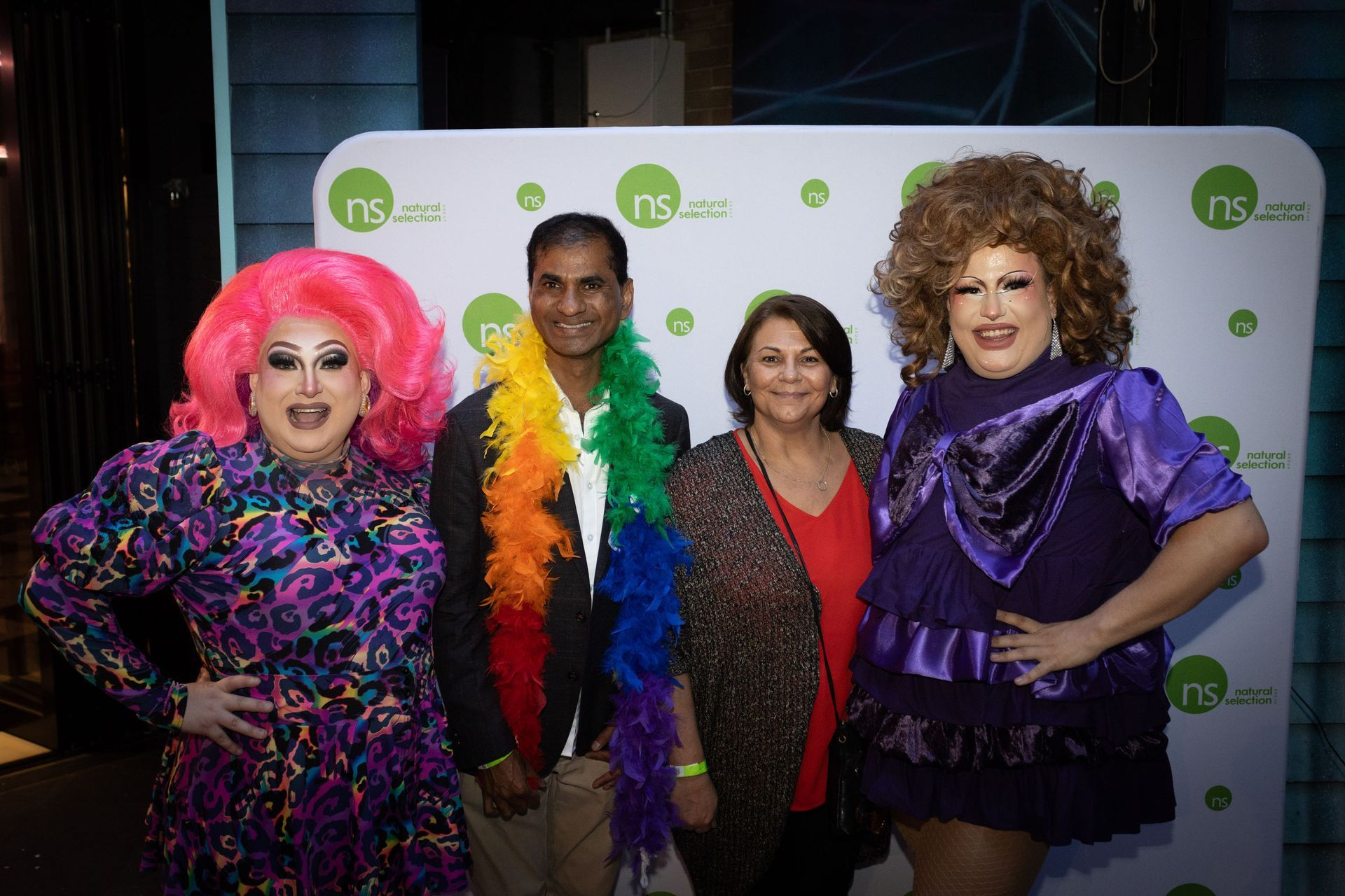Four people pose in front of a branded step-and-repeat backdrop, including two drag performers in vibrant costumes.