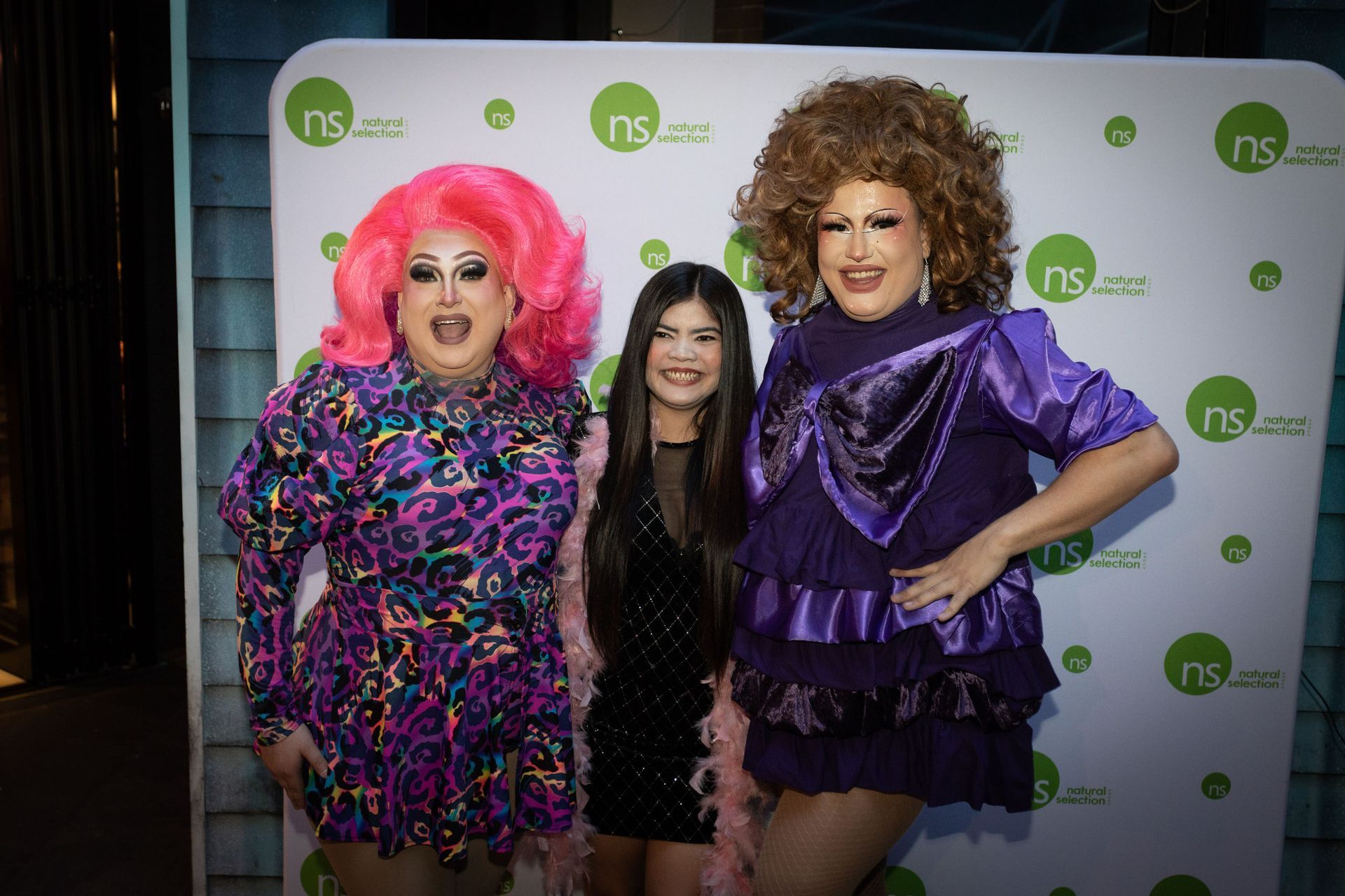 Three people stand in front of a white branded backdrop. Two are in vibrant drag outfits; one wears a black dress.