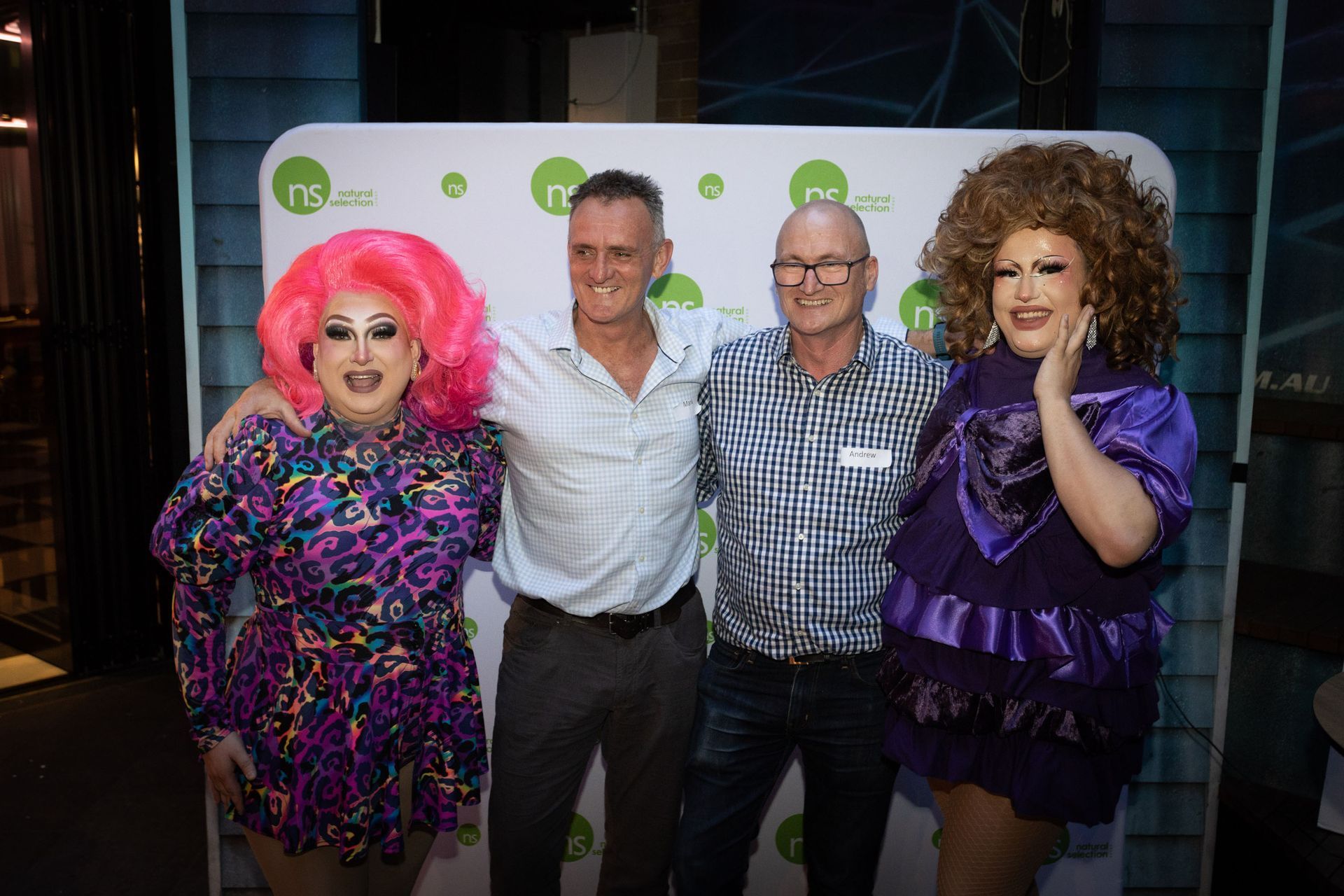 Four people pose in front of a branded backdrop, two in drag with elaborate hair and clothing, flanking two men in shirts.