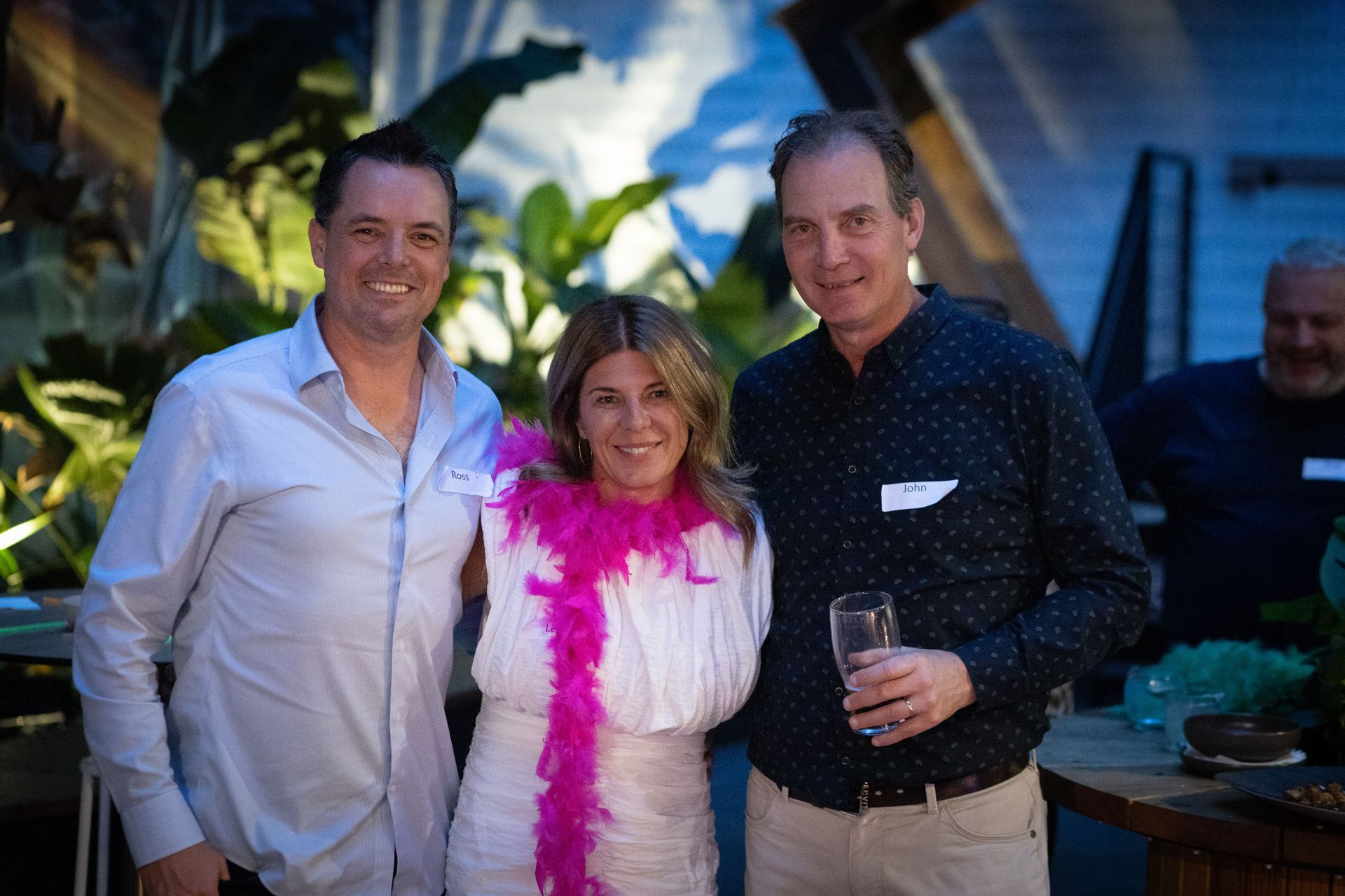 Three people pose together outdoors, smiling. One wears a pink feather boa, while the others hold drinks in a social setting.