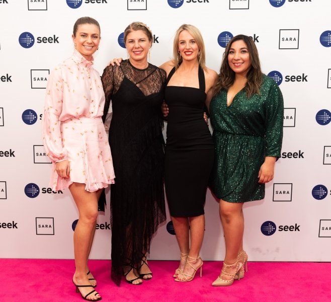 Four smiling people pose on a pink carpet in front of a white step-and-repeat backdrop with logos.