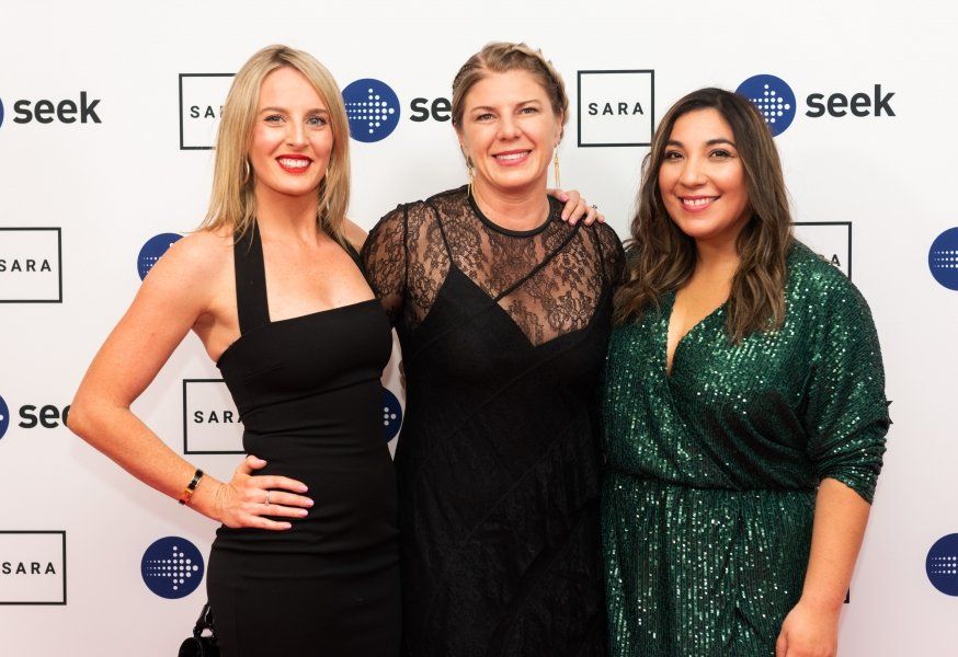 Three women smiling in front of a backdrop featuring the SARA and Seek logos at an event.