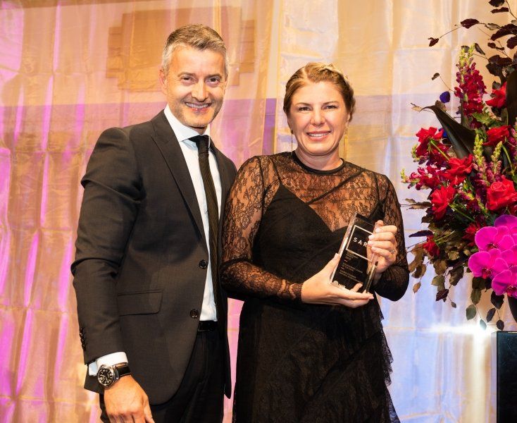 A smiling man in a suit and a woman in a black lace dress holding an award stand together at an indoor event.