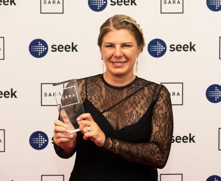 A smiling person in a black lace dress holds a glass award in front of a backdrop featuring 