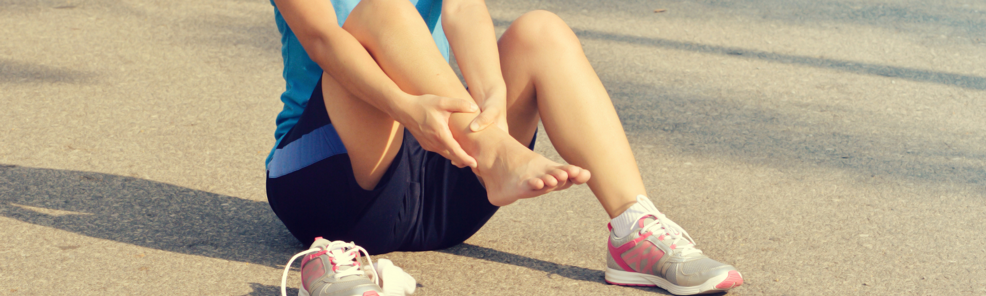 Runner seated on pavement, holding injured ankle beside running shoes at the roadside