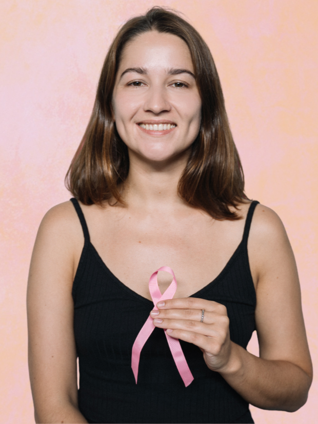 Smiling person in a black top holding a pink awareness ribbon against a peach background