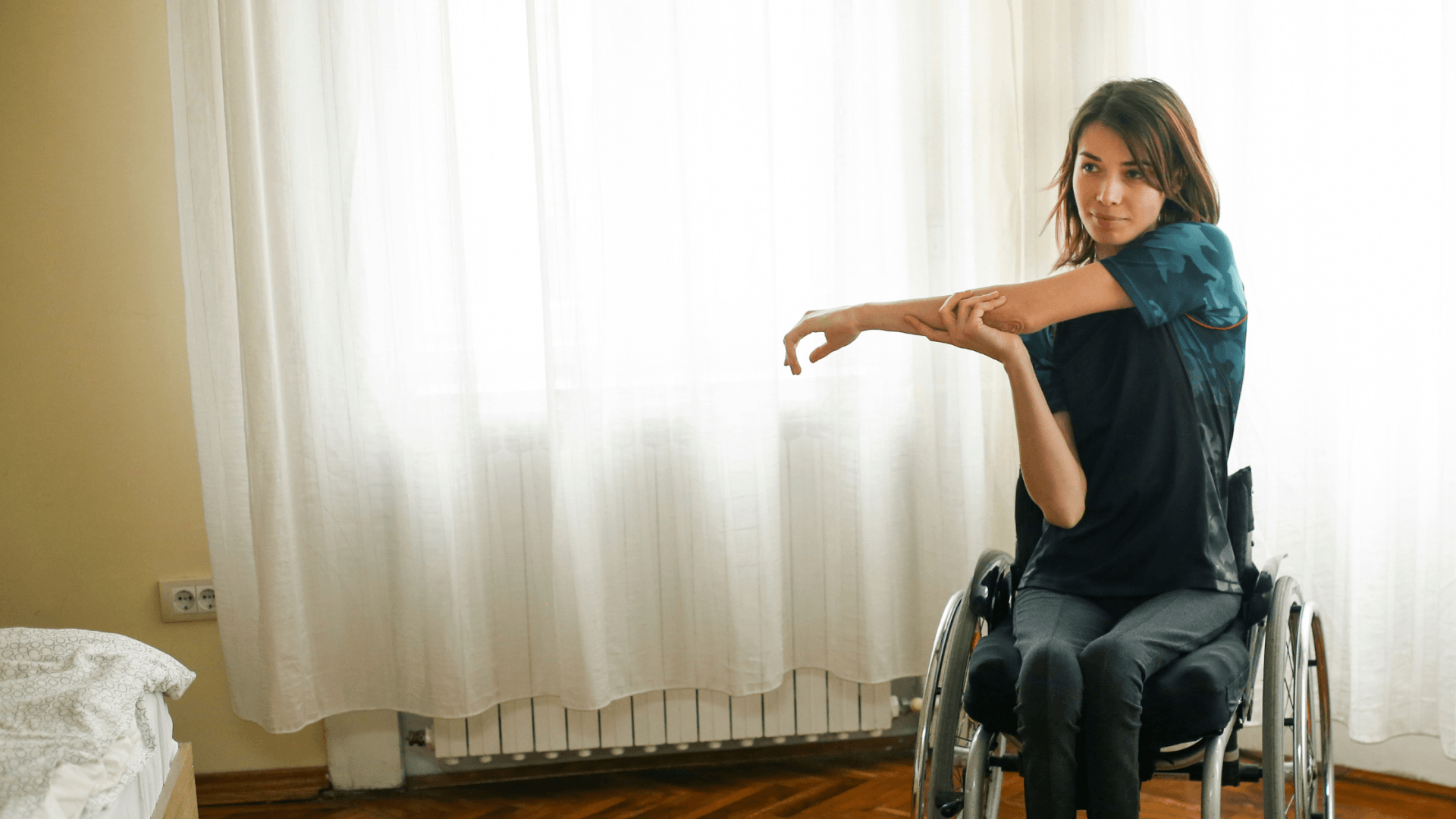 Woman in a wheelchair stretching her arm beside a bright window with white curtains