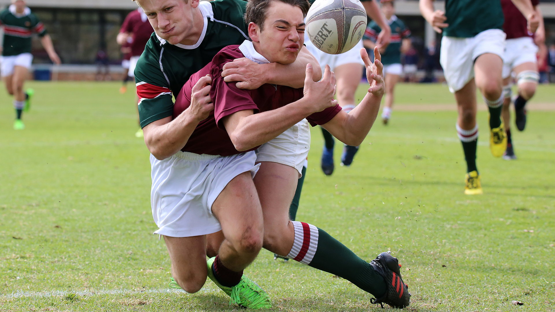 Rugby player in maroon and white tackles an opponent holding the ball on a grassy field.