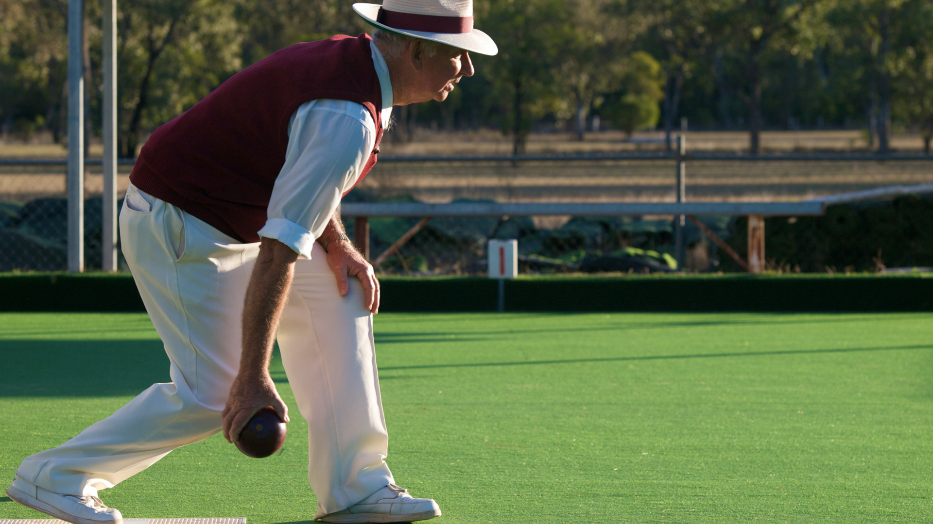 Bowler in white pants and burgundy vest crouches on a green lawn, preparing to roll a ball.
