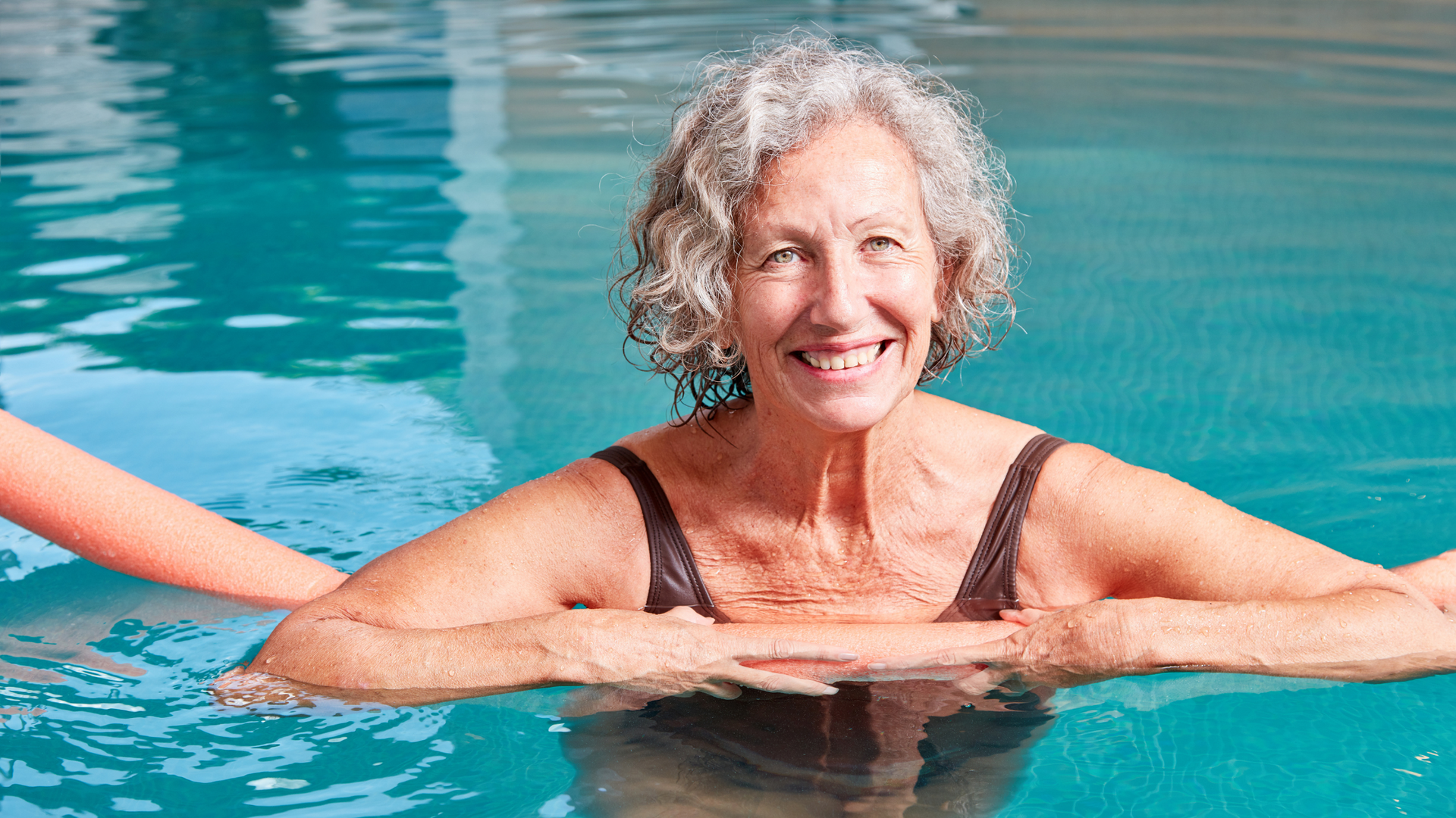 Smiling swimmer resting at the edge of a turquoise pool with an arm supporting her from behind