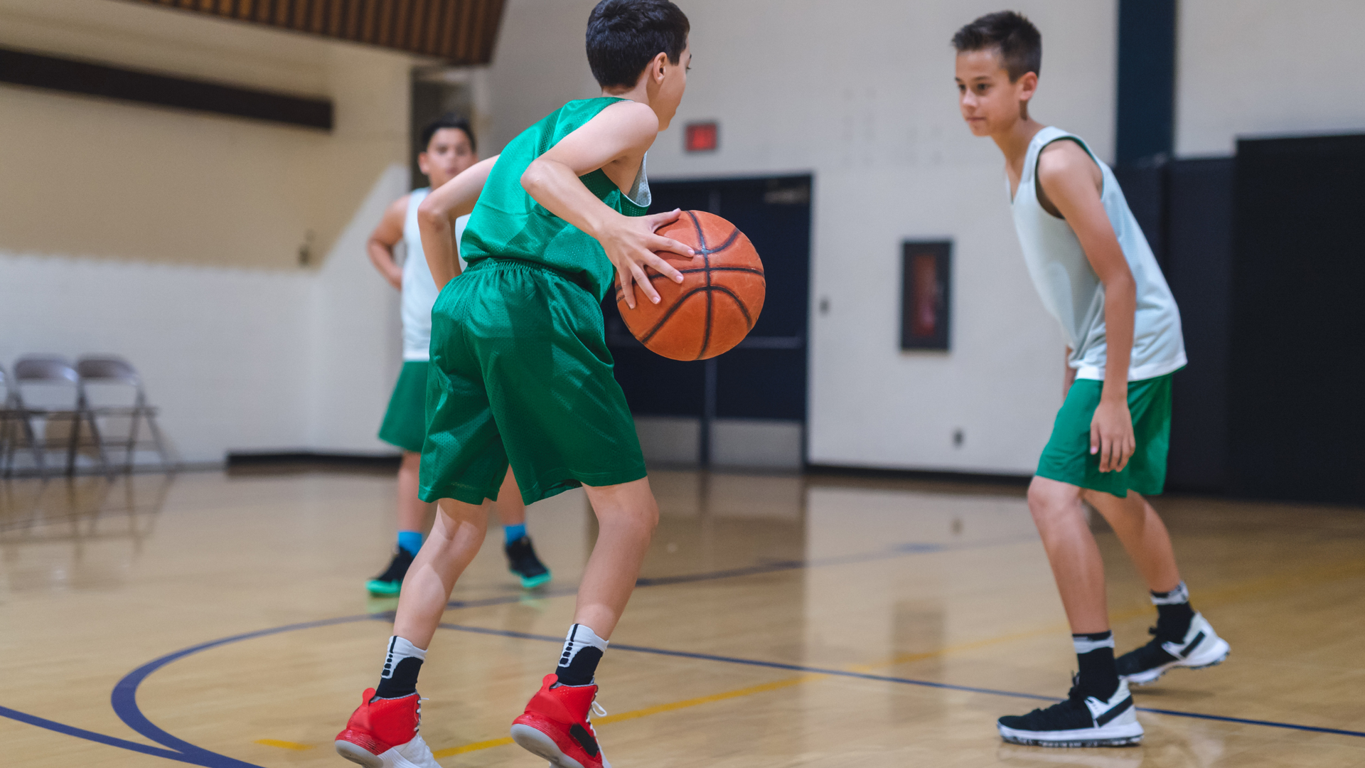 Kids playing basketball on an indoor court, with one dribbling while another defends.