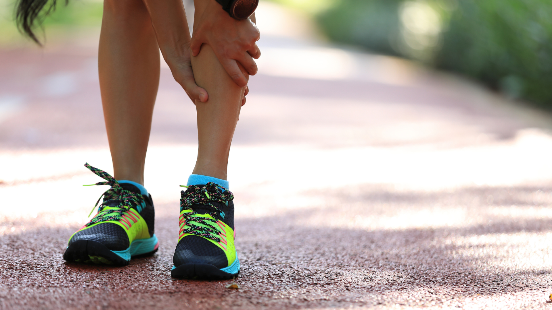 Runner stretching an ankle on a sunlit path, wearing black and neon green shoes.