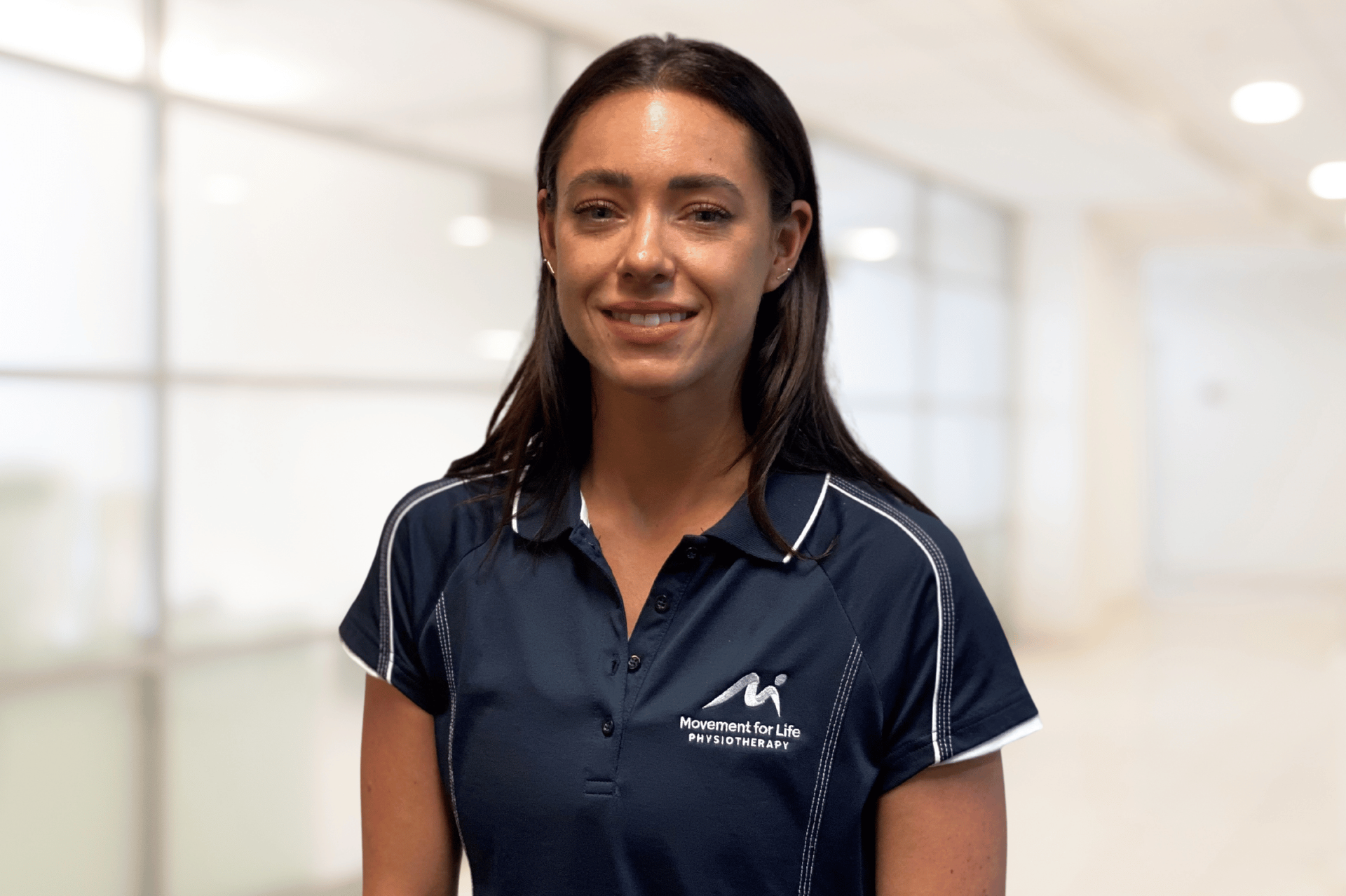 Woman in a navy polo shirt standing indoors against a bright blurred background.