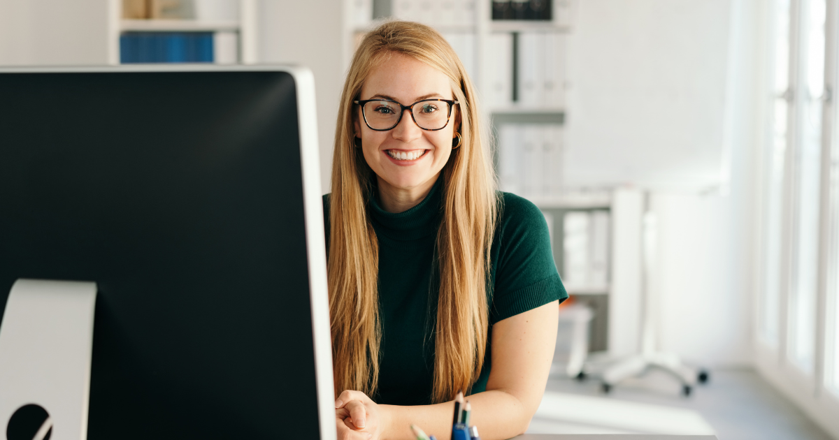 Smiling person with long blond hair at a desk, partially hidden by a large monitor in a bright office