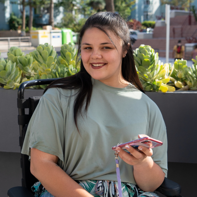 Smiling person in a light green shirt holding a phone outdoors near potted succulents