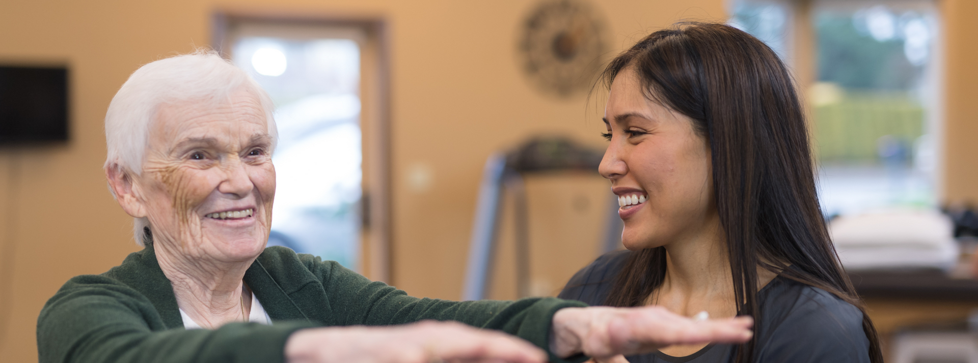 Smiling woman and elderly person chatting indoors, both leaning on a table