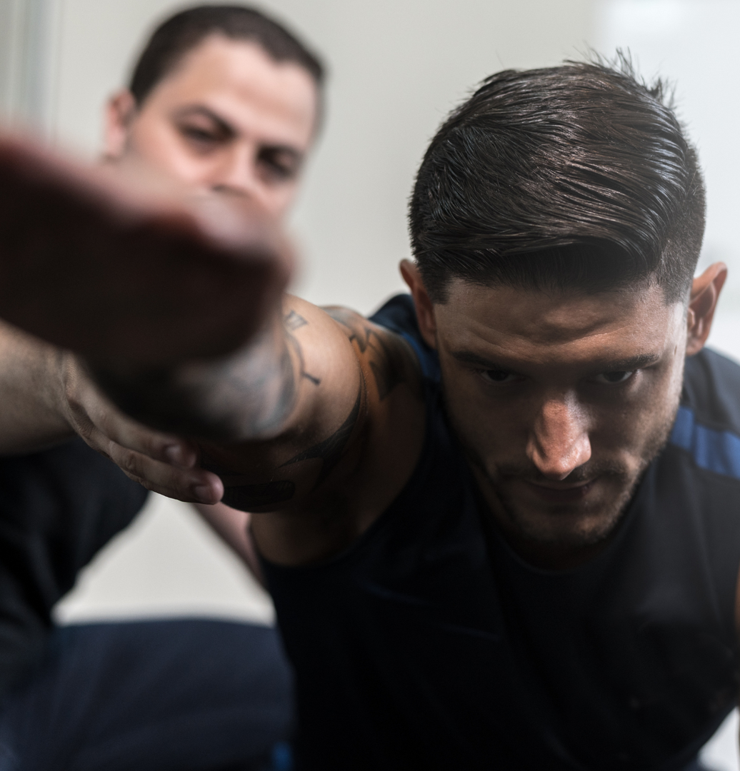 Man in a gym leaning forward with an outstretched arm, focused, with another person blurred in the background