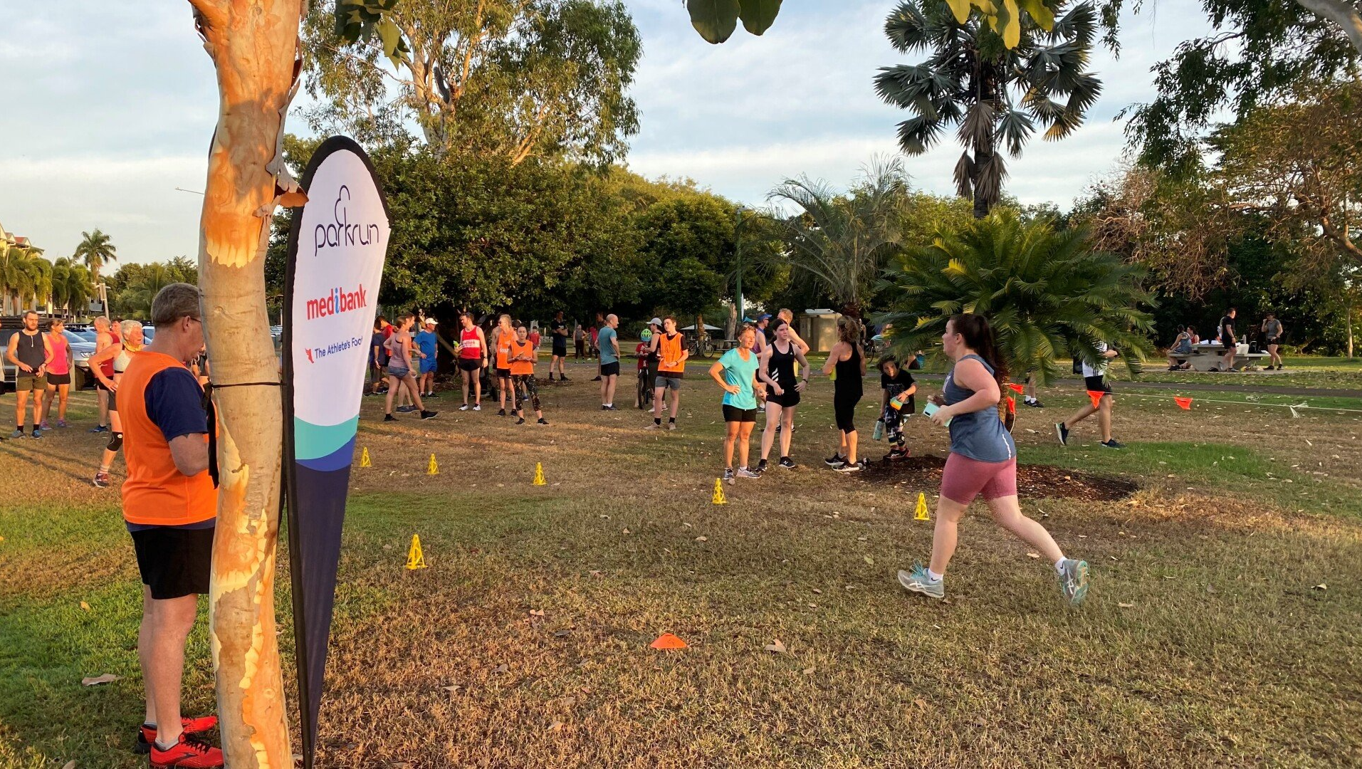 People gathered outdoors in a grassy park for a running event, with volunteers and a finish flag nearby.