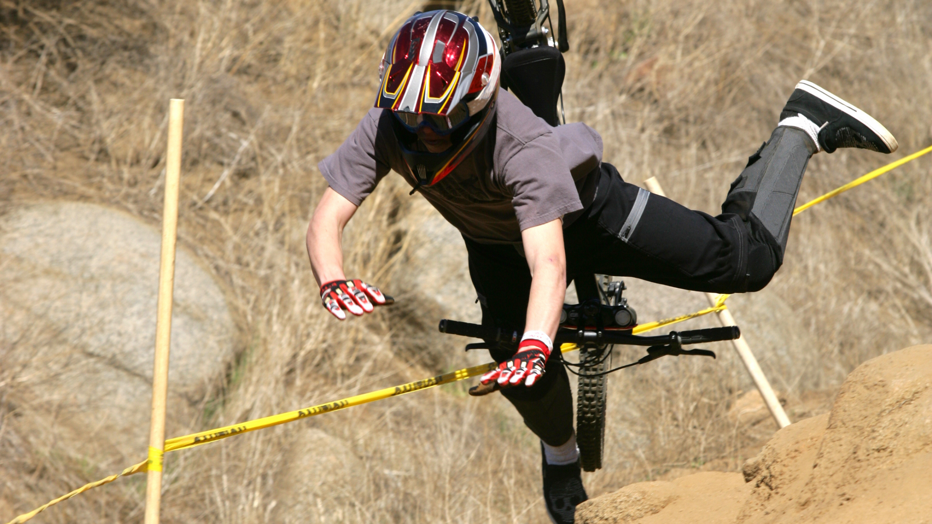 Mountain biker crashing over a dirt jump, legs up, helmet on, beside yellow course tape.