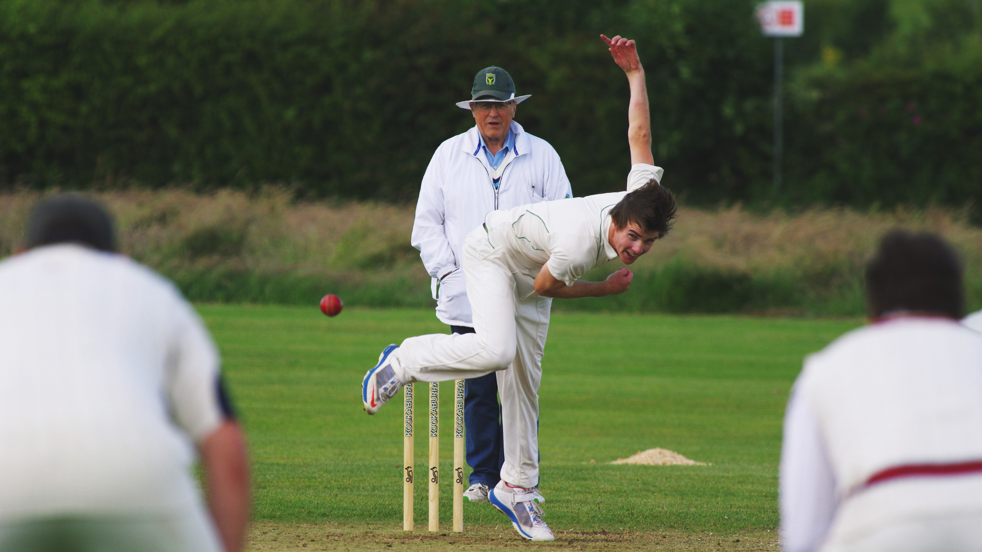 Cricket bowler in white delivers a ball toward the stumps on a grassy field while fielders watch.