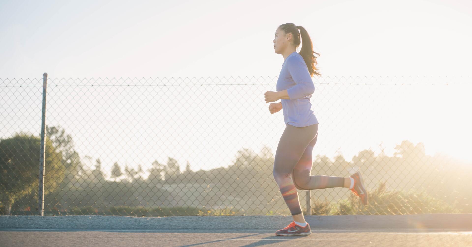 Woman jogging on an outdoor track at sunset, wearing a blue shirt and black shorts near a chain-link fence.