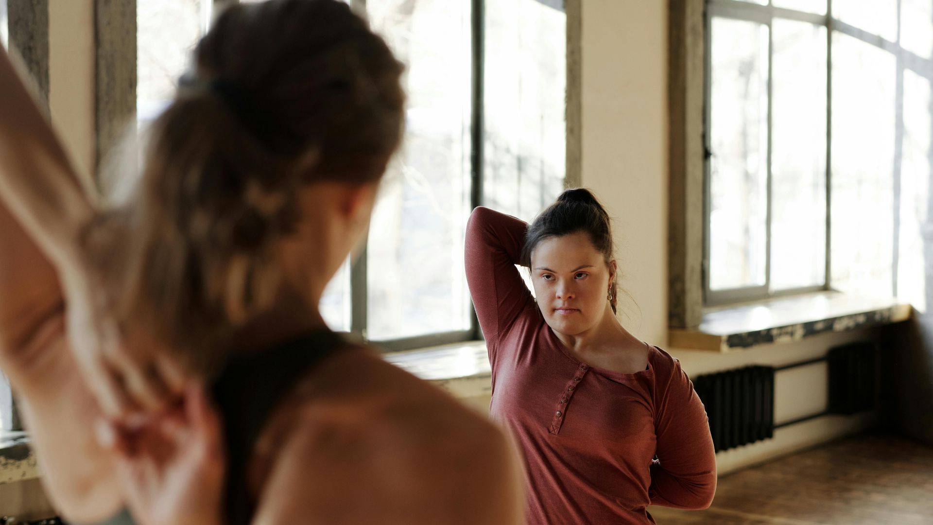 Two people stretching in a sunlit studio with large windows and hardwood floors