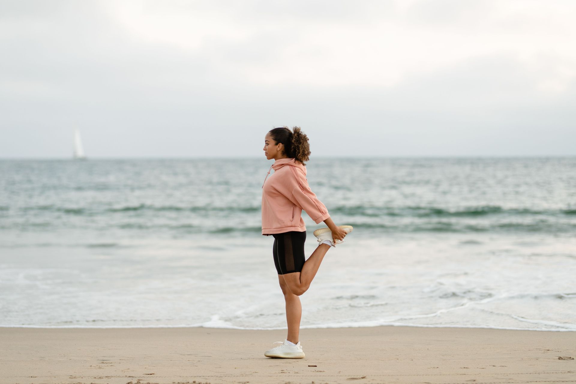 Person stretching on a beach, standing in profile near the shoreline under a cloudy sky.