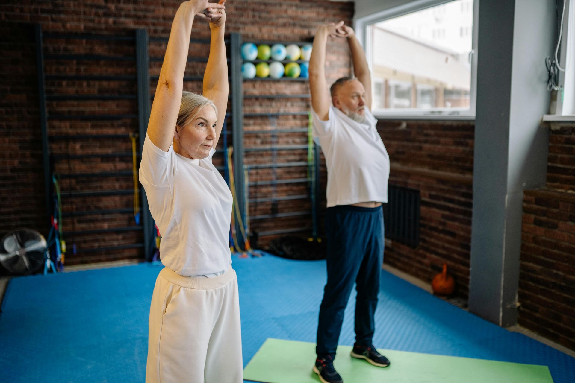 Two people stretching overhead in a gym with blue mats and brick walls