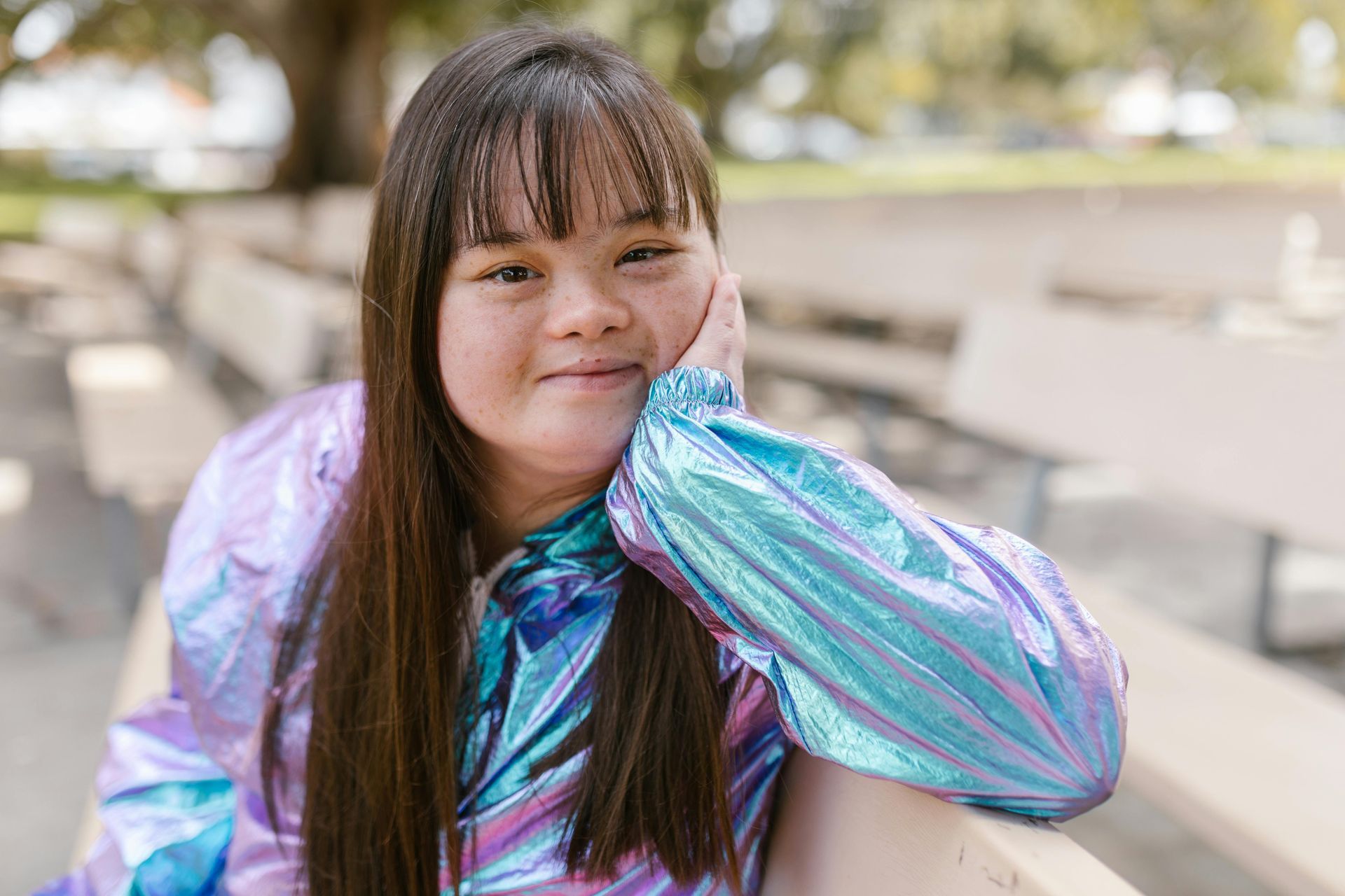 Person in a shiny blue jacket leaning on a bench outdoors, smiling softly.
