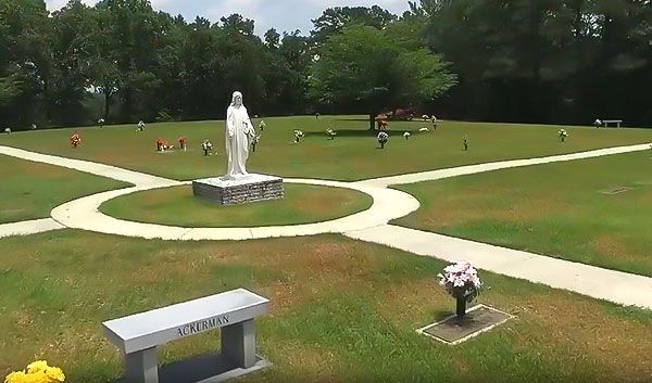 Cemetery with a statue of Mary at the center, surrounded by graves and pathways, a bench in the foreground.