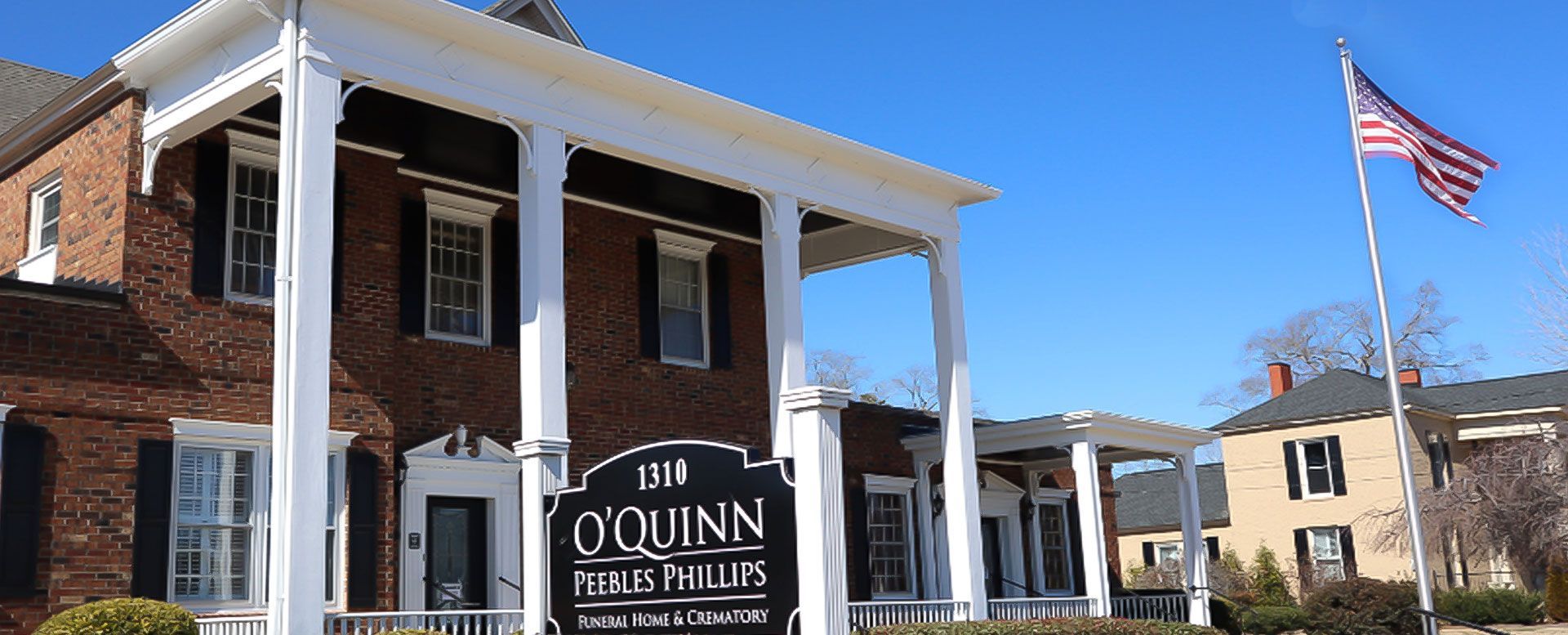 Brick building with white columns, O'Quinn Funeral Home sign, American flag waving in the blue sky.